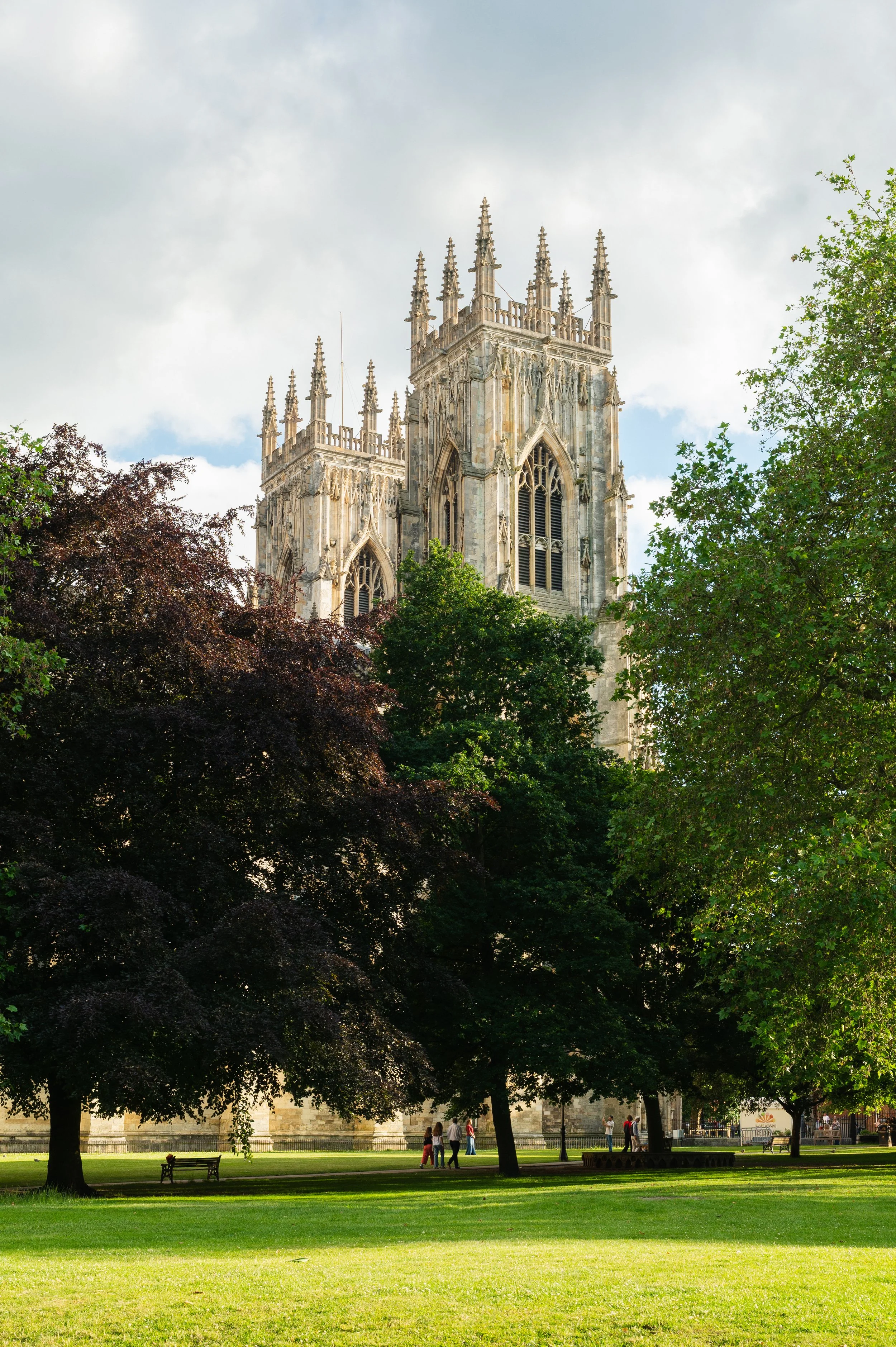 View of a historic church tower with Gothic architecture, partially obscured by lush green trees, with people walking in a park area.