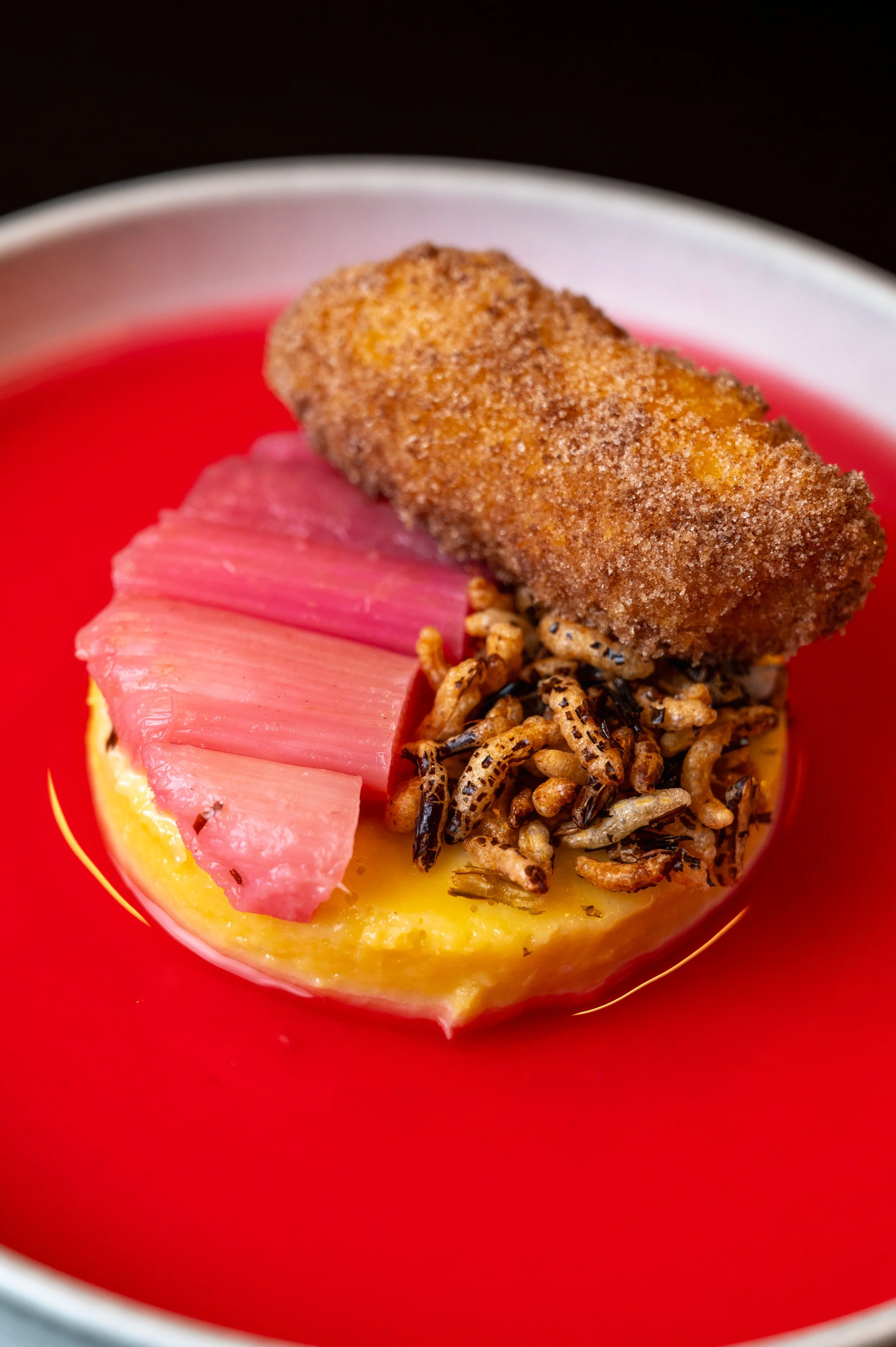 Close-up of a small dessert in a red cup, topped with a breaded item, pink gelatinous pieces, crispy grains, and a yellow base, possibly custard or pudding, with a dark background.