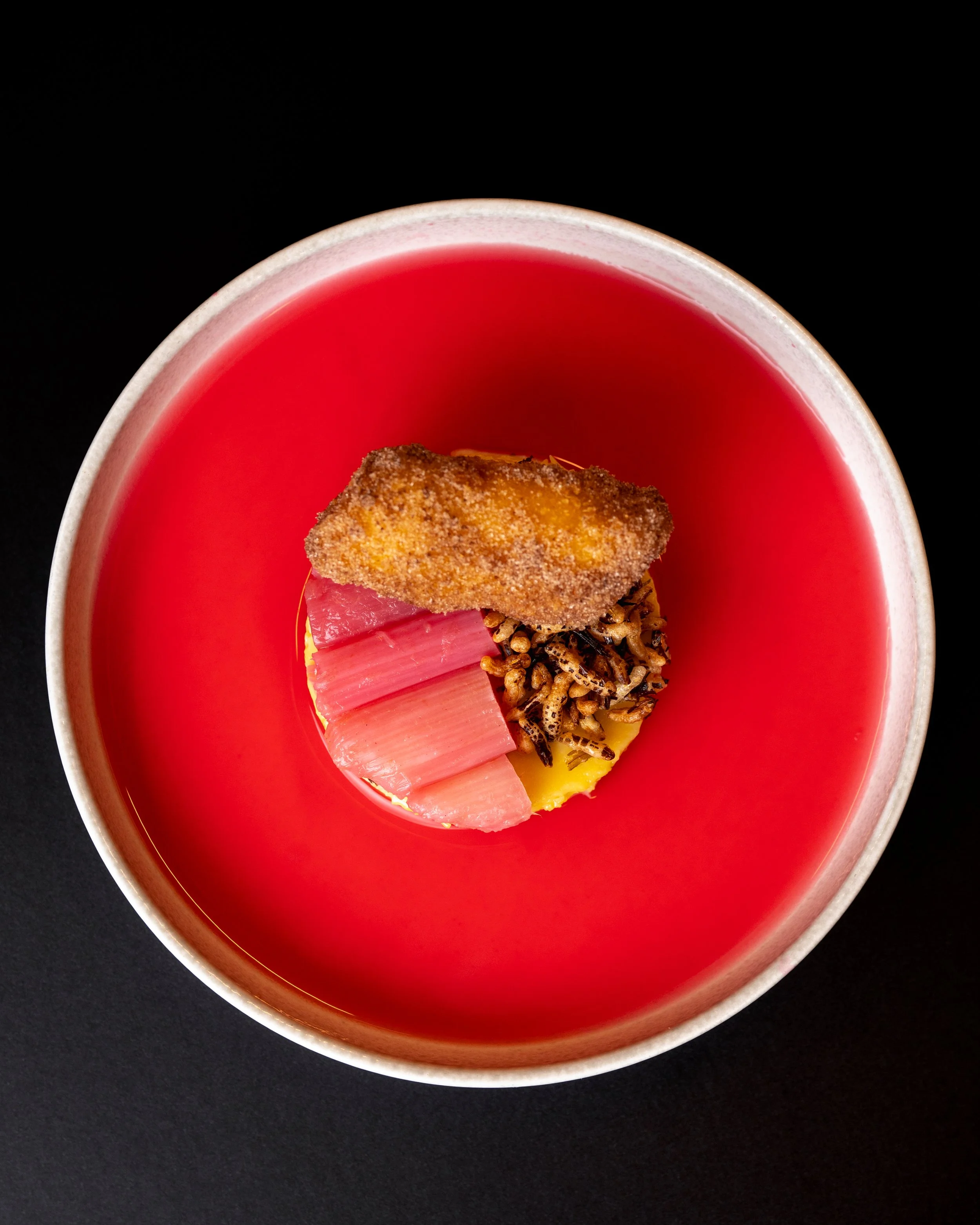 Close-up of a red bowl of vibrant liquid with a piece of breaded fried food and some grains or seeds inside, on a black surface.