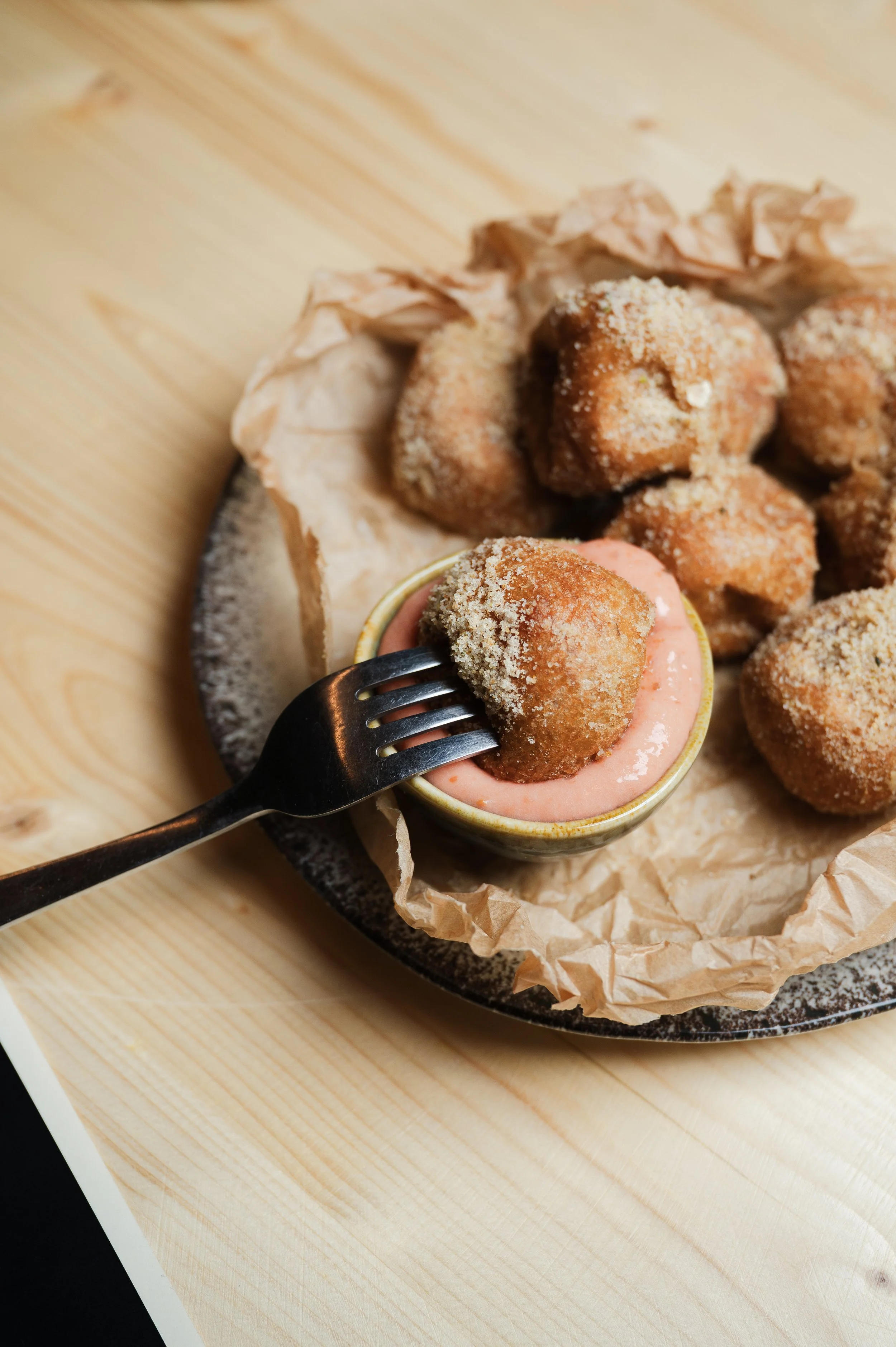 Close-up of bite-sized fried food balls covered in crumbs, served in a small cup with pink dipping sauce, on a black plate with parchment paper, on a light wooden surface.