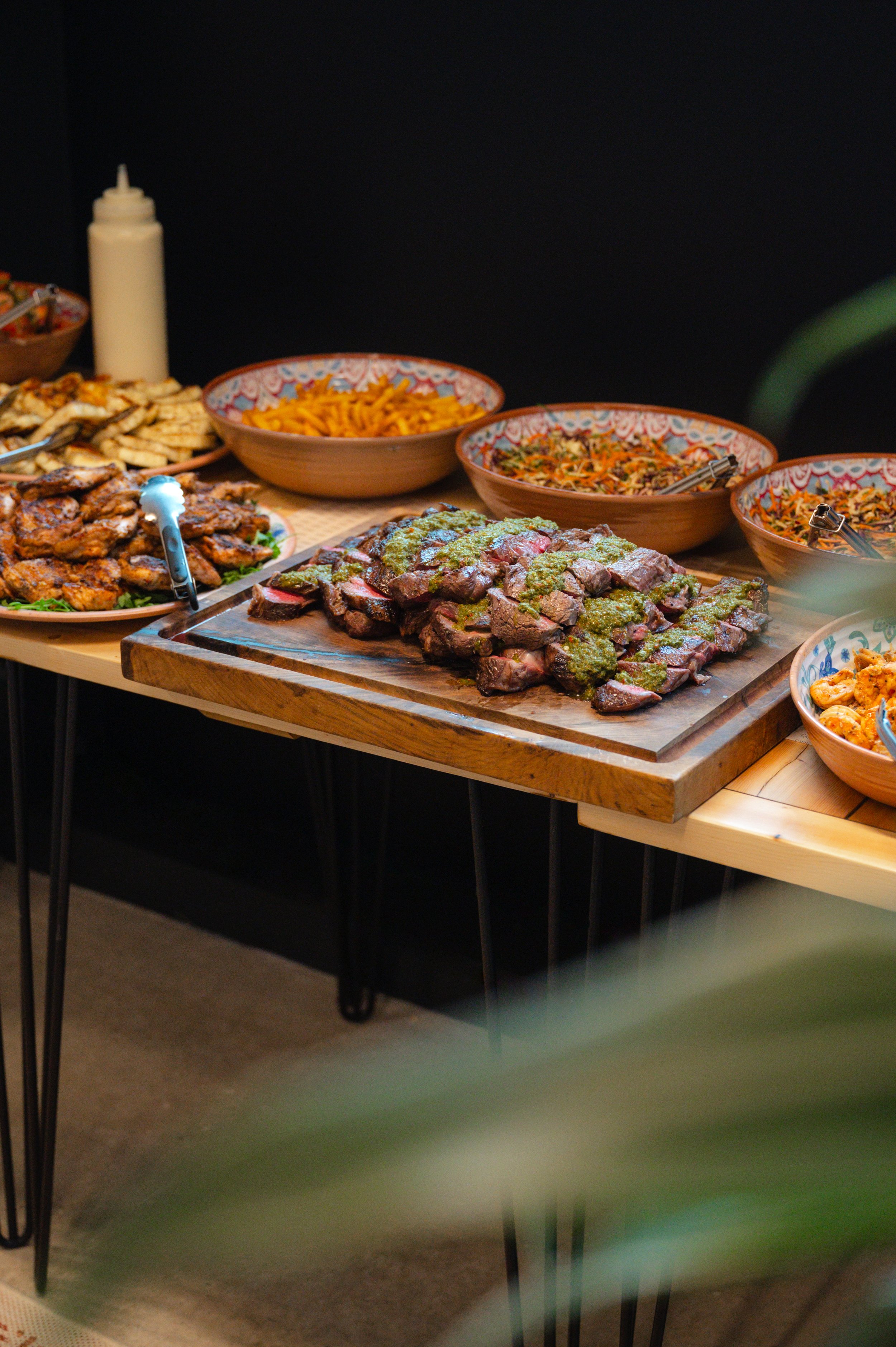A table with various dishes including sliced beef with sauce, pasta, grilled chicken wings, and salads in colorful bowls, set against a dark background.