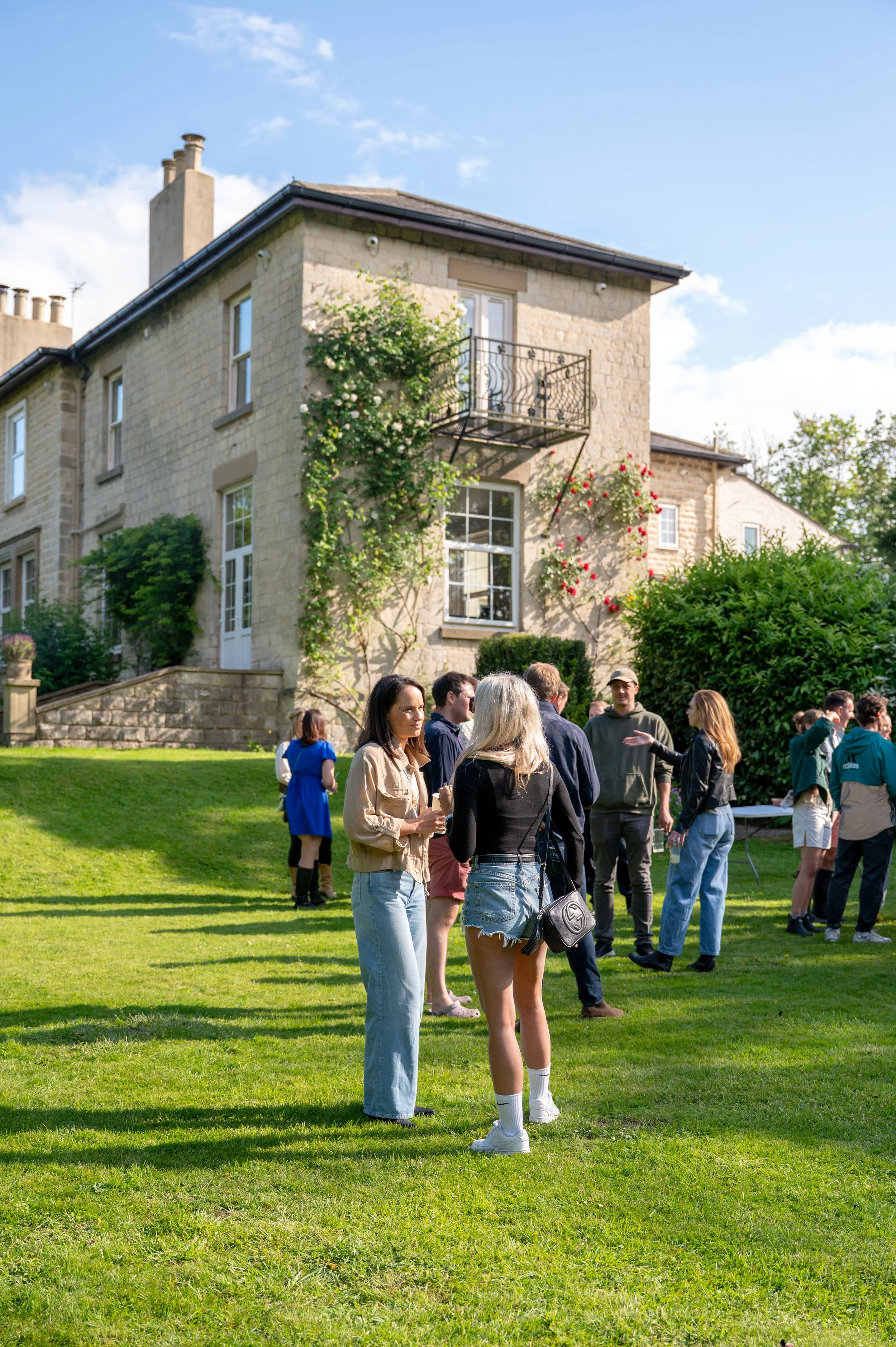 A group of people gathered on a lush green lawn in front of a beige stone house with climbing plants and colorful flowers. The sky is blue with scattered clouds.