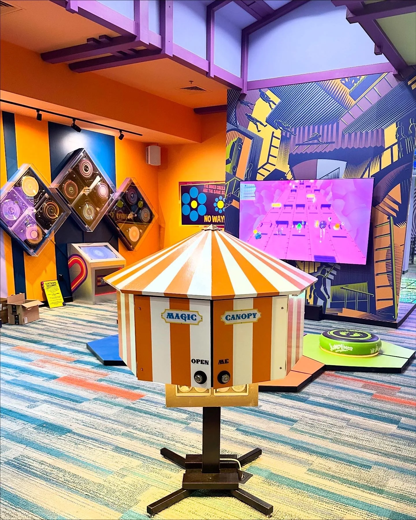 An indoor arcade featuring a spinning carnival-style game with a striped orange and white canopy labeled "Magic Canopy," a retro-style colorful mural, and large screen displays showcasing arcade games.