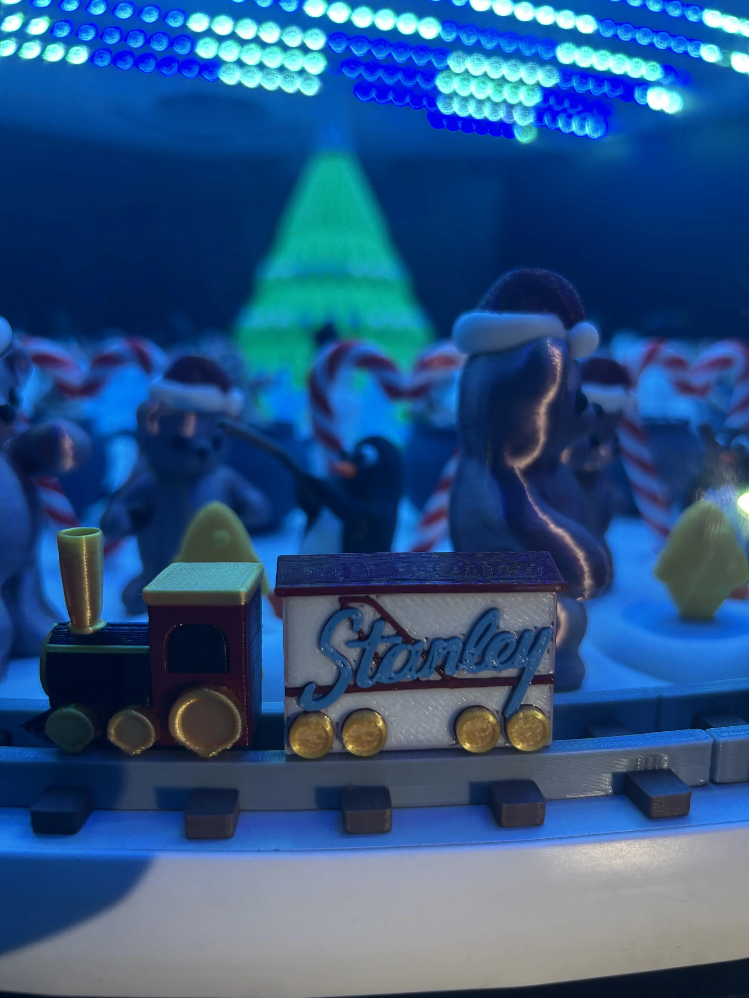 Close-up of a miniature toy train with the word 'Stanley' on its side, placed on a track with blurred festive holiday scene in the background, including people wearing Santa hats and a decorated Christmas tree under blue holiday lights.