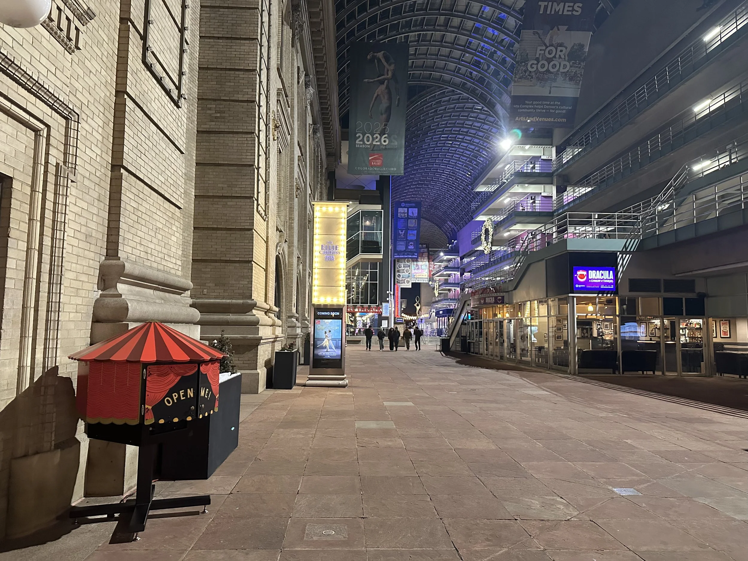 Empty shopping area with a small red and black puppet theater booth labeled 'Open Me!' on the left, with a historic brick building behind it, and modern multi-story buildings with illuminated signs and banners on the right, under a curved glass ceili