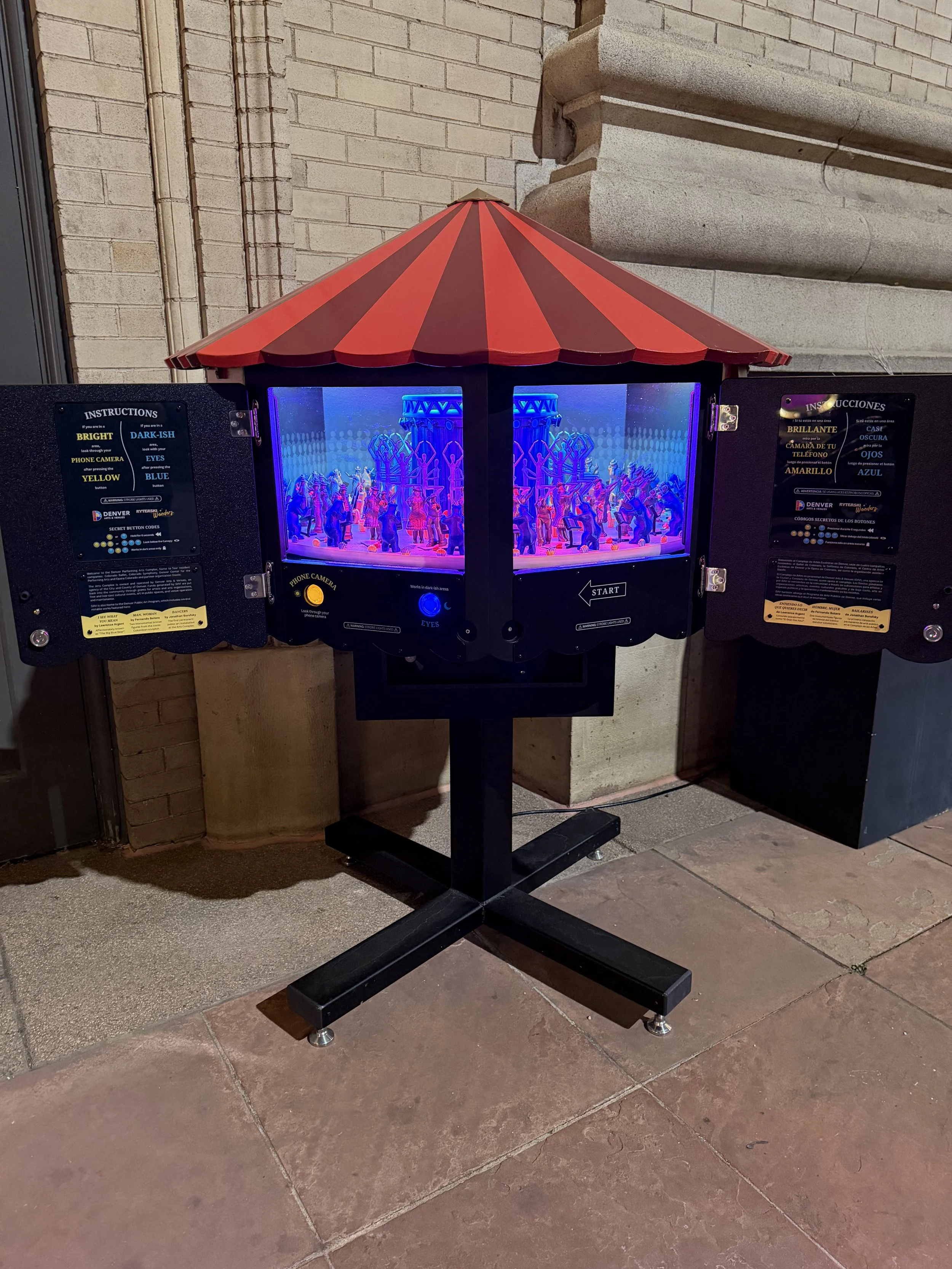 Miniature carousel photo booth with a red and dark brown striped roof, illuminated by purple and blue lights, with a play button and instructions on the sides.