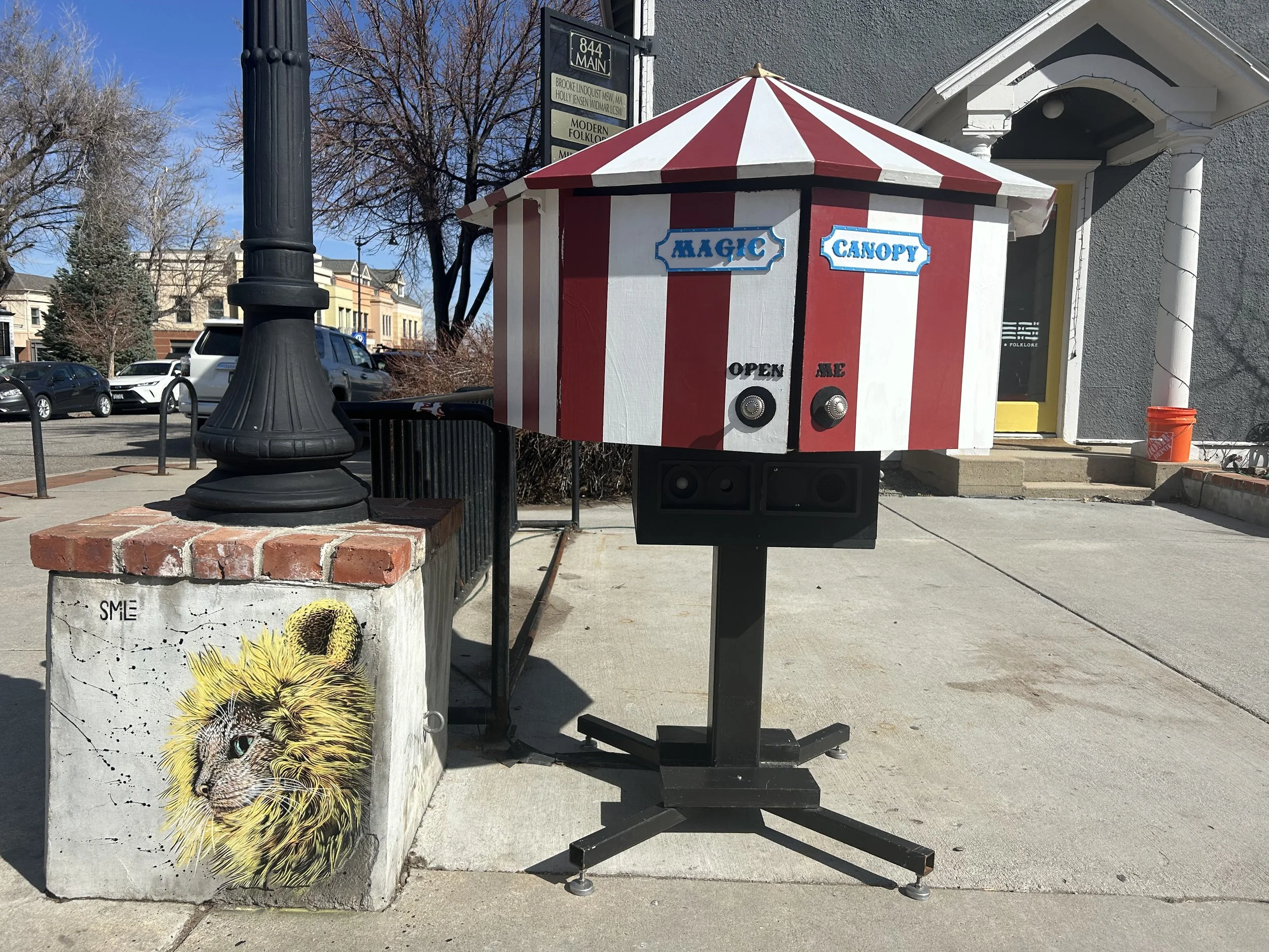A street corner with a painted concrete block featuring a lion's face and the word 'SMILE,' a black lamppost on a brick base, and a striped red and white carnival-style booth labeled 'Magic Canopy' with buttons and speaker equipment.