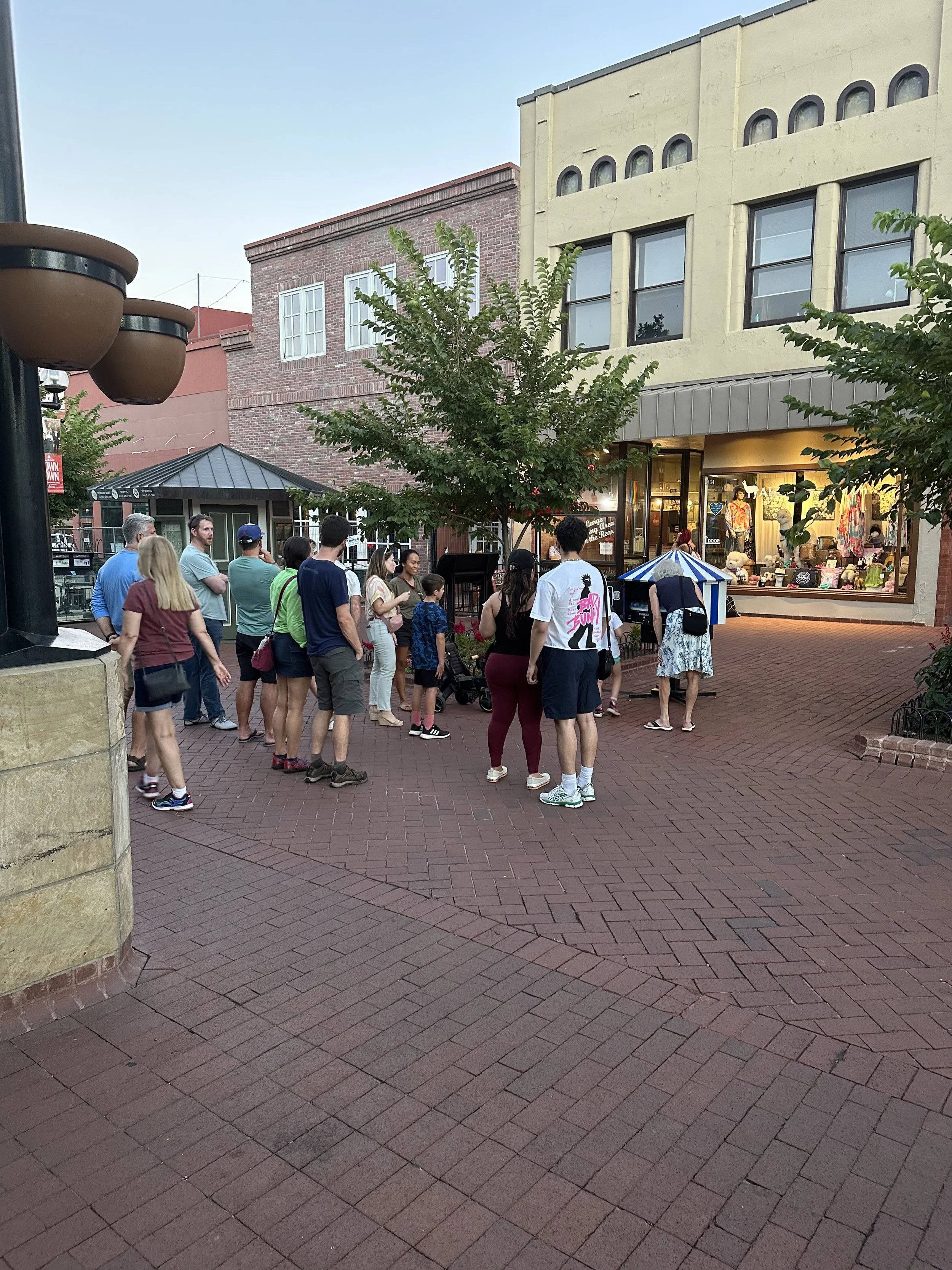 People gathered outside a storefront with large windows on a brick-paved sidewalk, with some trees and buildings in the background.