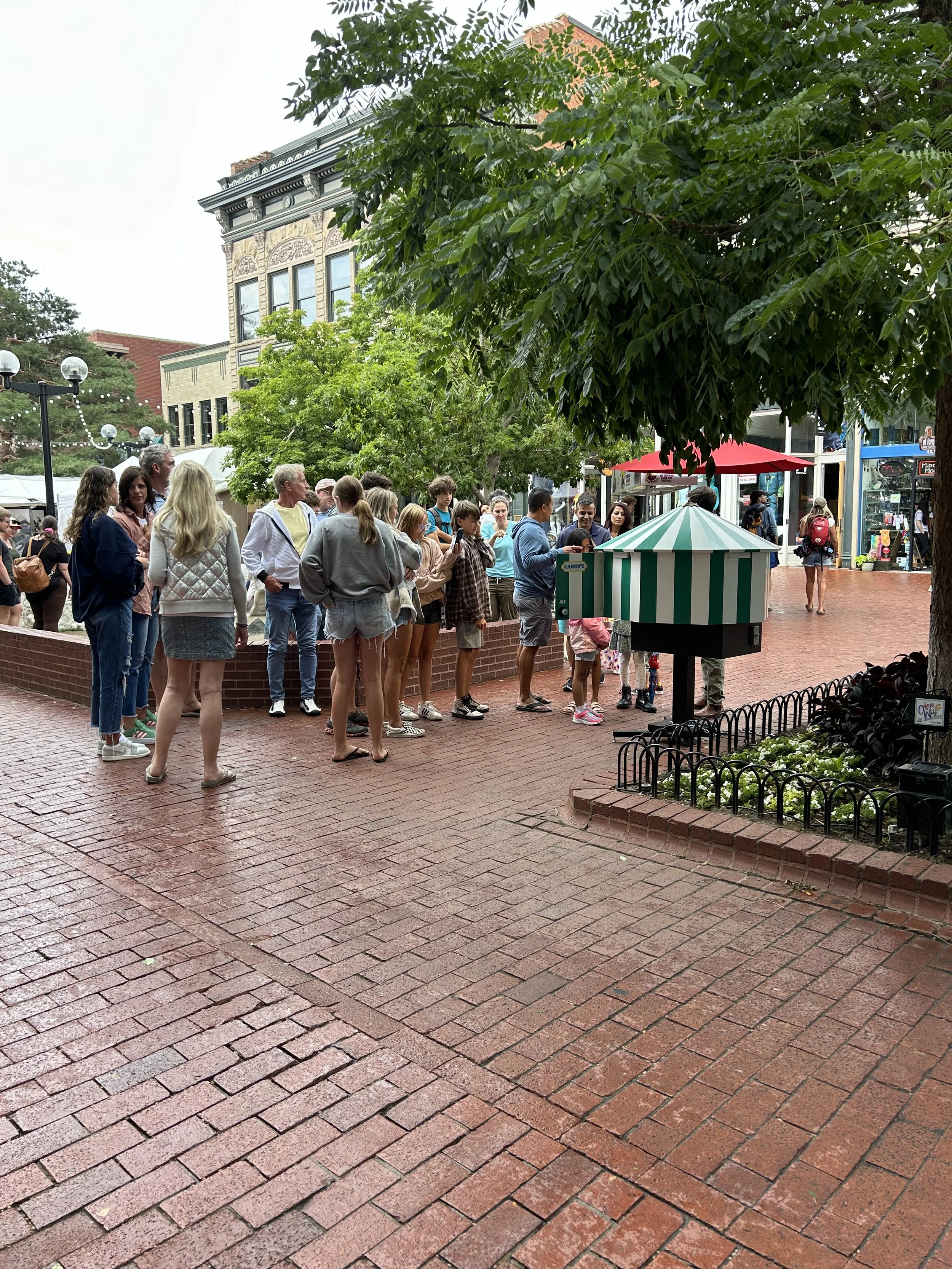 Group of people standing in a line outdoors on a brick sidewalk with shops and trees in the background.