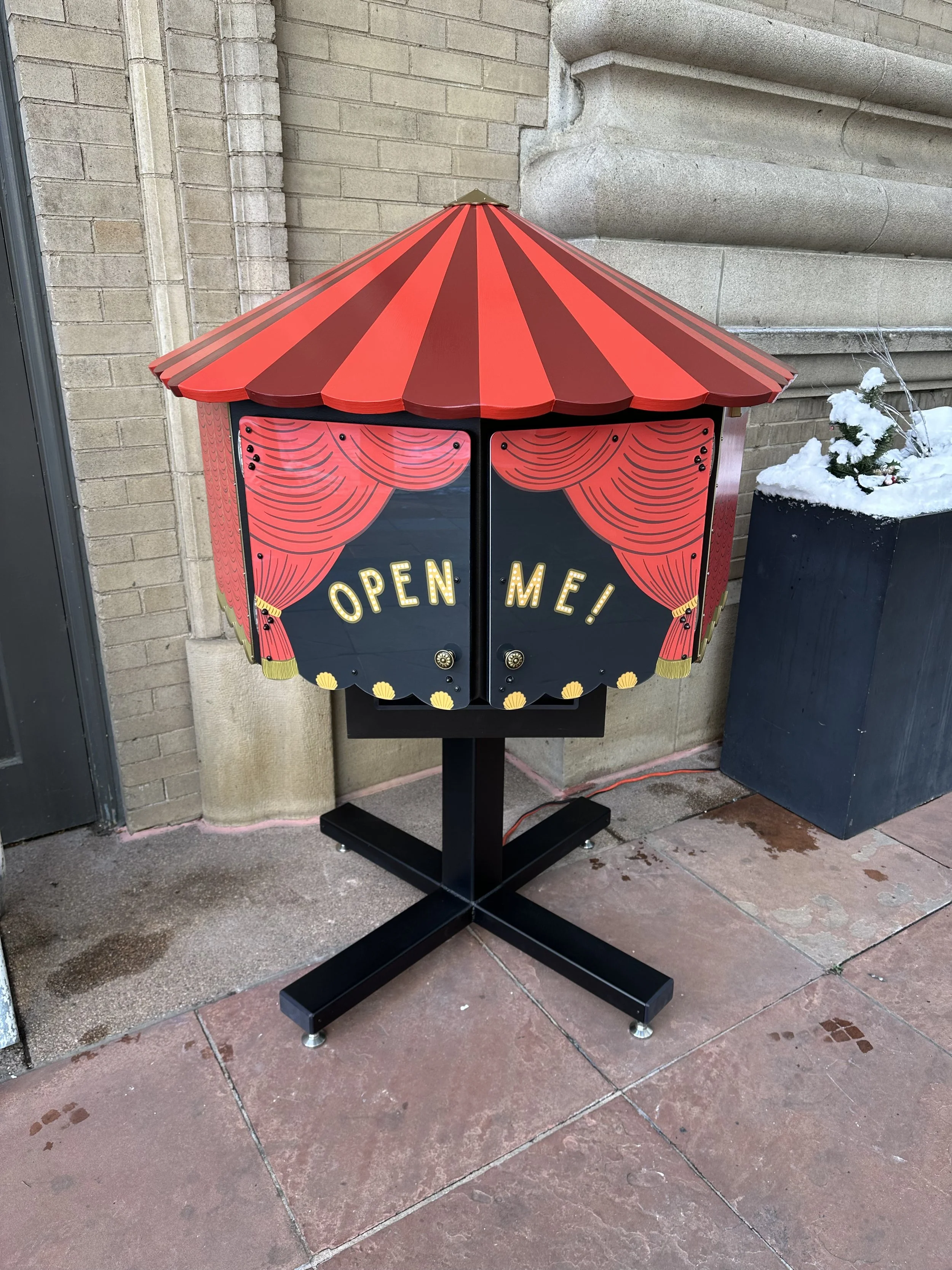 A circus-themed sidewalk sign with a red and black striped canopy design and red curtains, displaying the words "OPEN ME!" in gold letters.