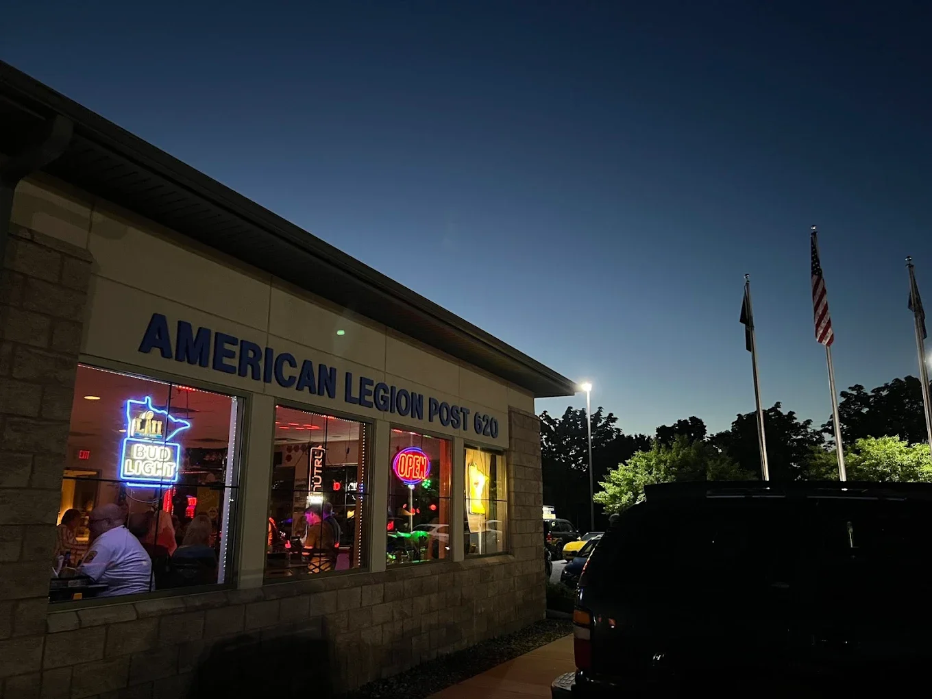 Nighttime view of American Legion Post 620 with illuminated neon signs inside the building and flags flying outside, with trees and cars in the background.