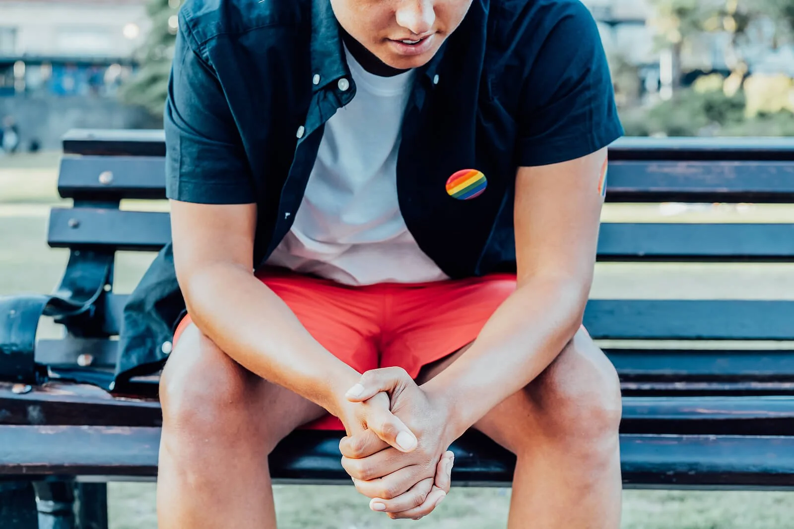 a person with a rainbow button sits on a park bench and looks sad