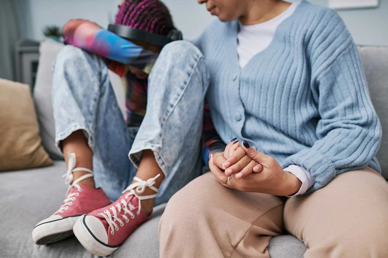 A mom holding the hand of her teen, who has pink hair and is crying