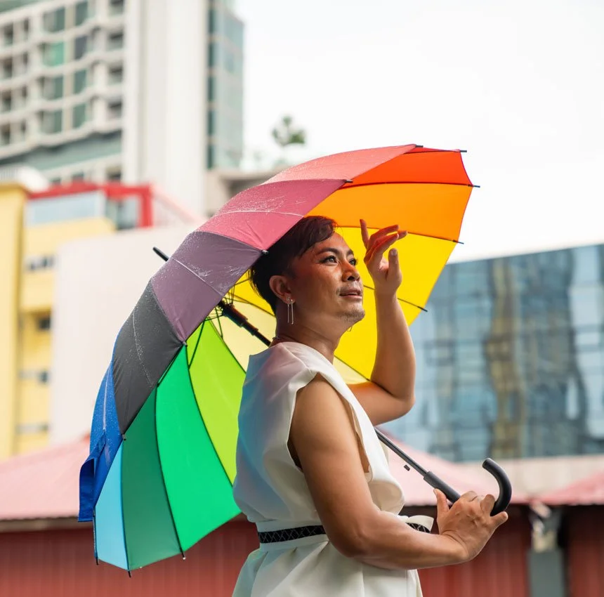 Drag queen in a white dress holding a rainbow umbrella