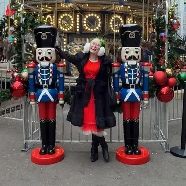 A silly woman wearing very large white earmuffs posing with two nutcrackers in front of a carousel