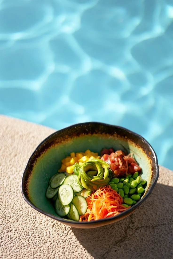 A colorful salad with cucumber slices, shredded carrots, edamame, corn, avocado, and diced tomato in a bowl near a swimming pool.