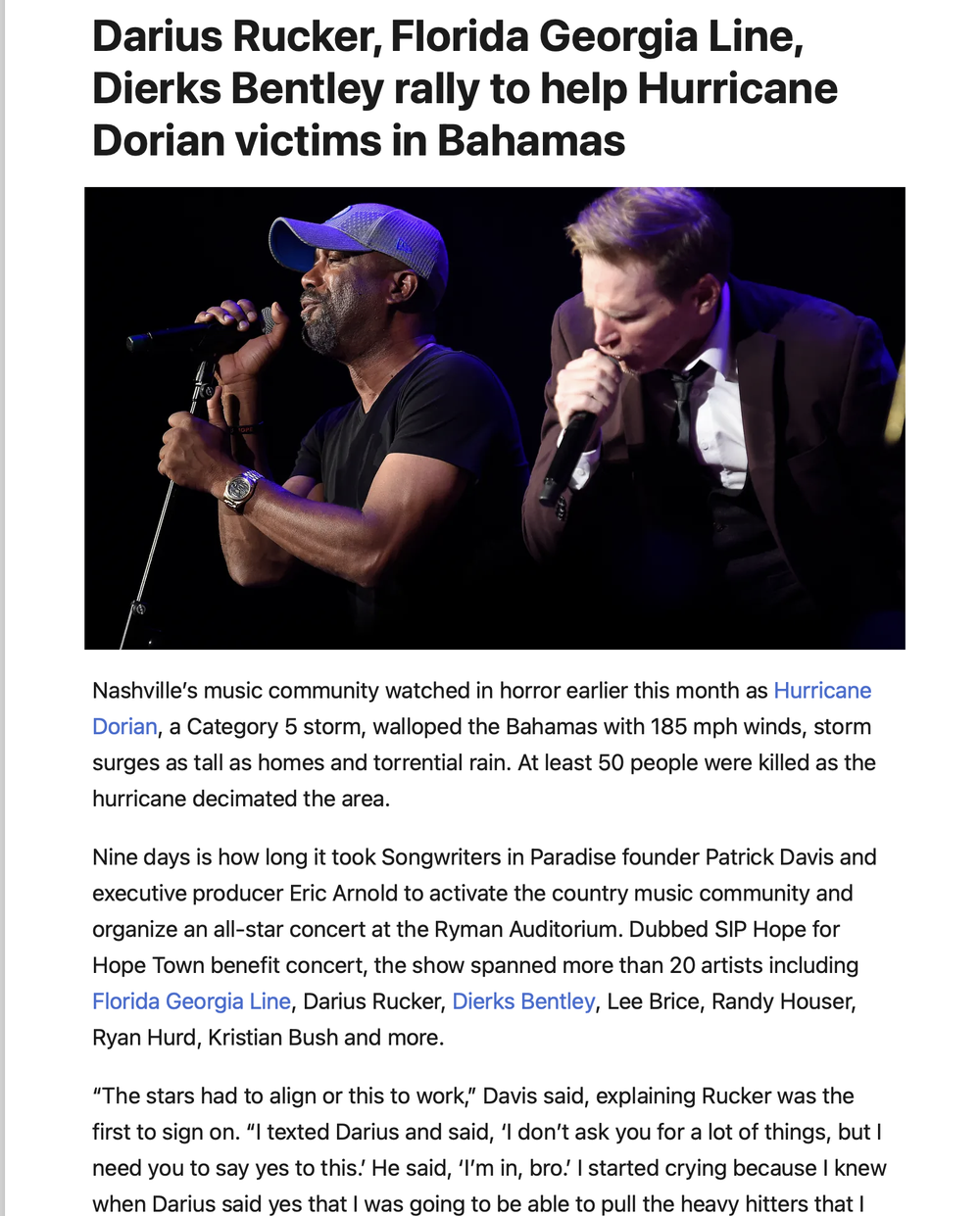 Two men on stage, one wearing a cap and black t-shirt, the other in a dark suit, both singing into microphones during a rally.