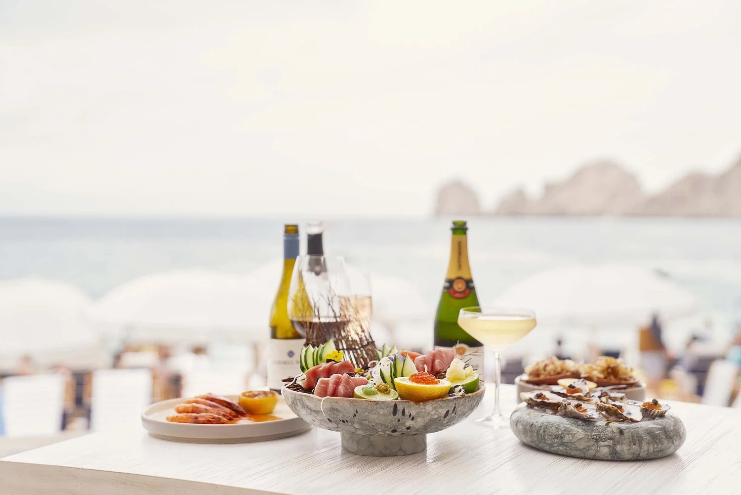Beachside table with assorted seafood, drinks, and a salad, overlooking the ocean with mountains in the background.