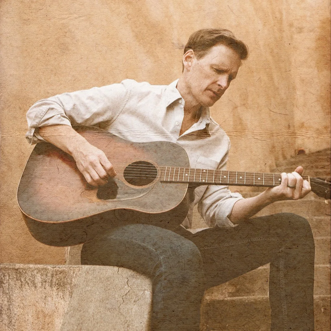 Patrick Davis sitting on a stone, playing an acoustic guitar with a wooden background