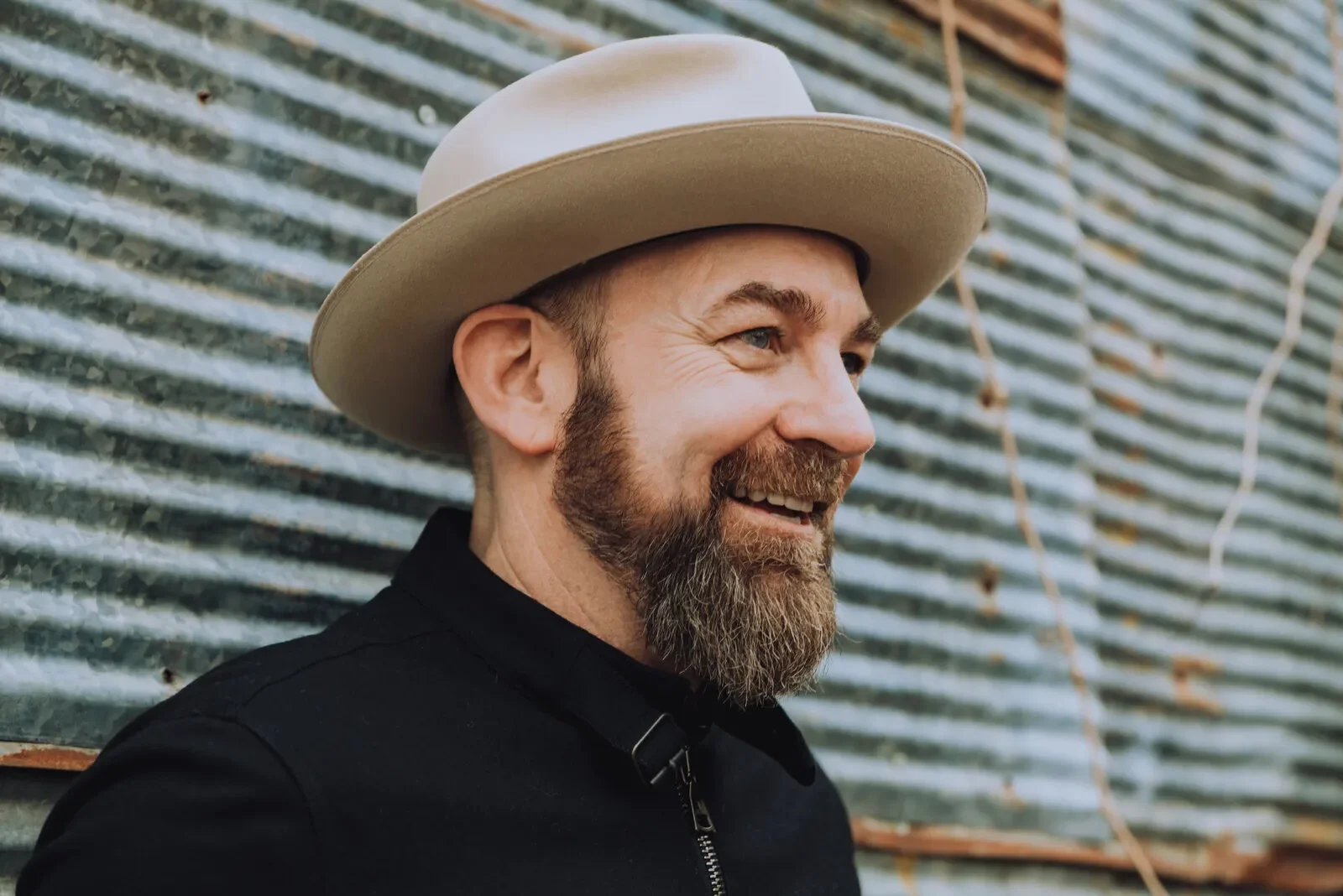 Kristian Bush with a beard wearing a beige wide-brimmed hat and black clothing standing in front of a corrugated metal background.