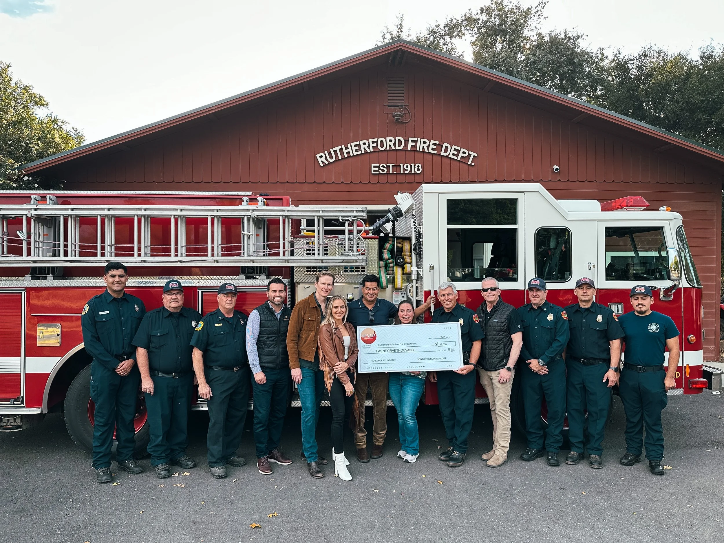 Group of people, including firefighters and civilians, standing in front of a fire truck and a fire station, holding a large donation check.