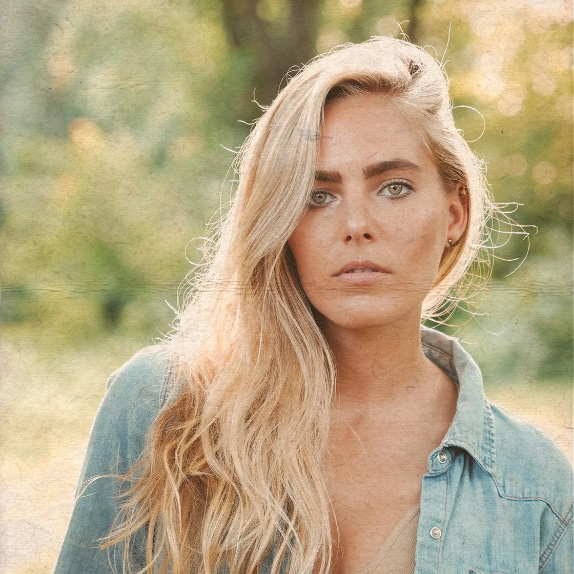 Lauren Jenkins with long, wavy blonde hair, wearing a denim shirt, outdoors with greenery in the background.