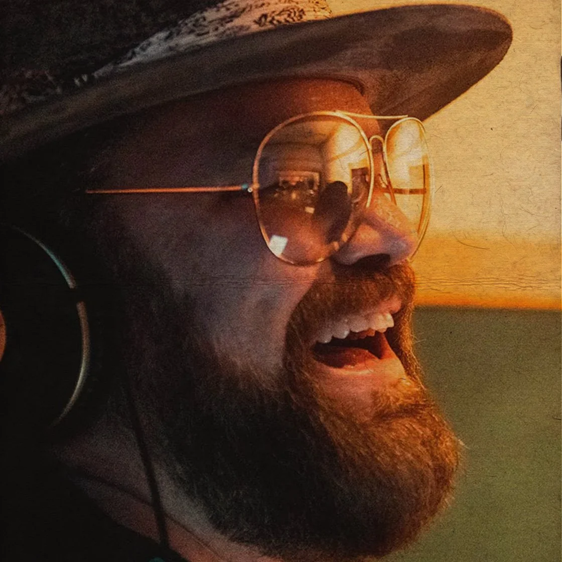 James Otto with a beard, wearing glasses and a hat, smiling and laughing, with a warm, orange-tinted background.