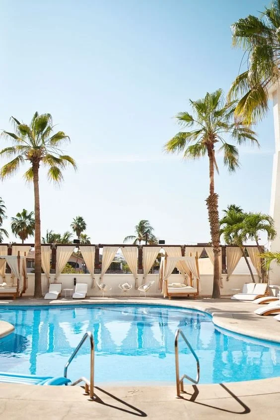 Swimming pool area with lounge chairs and cabanas surrounded by tall palm trees under a clear blue sky.