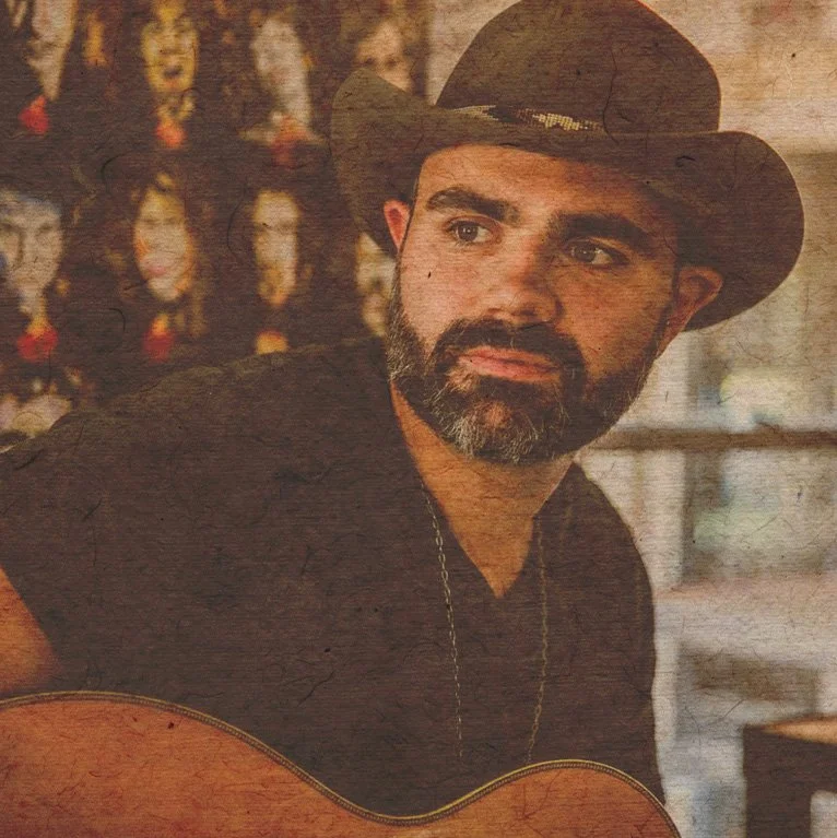 Django Walker with a beard wearing a black hat and black shirt, holding a guitar, looking to the side in a rustic setting with masks hanging on the wall in the background.