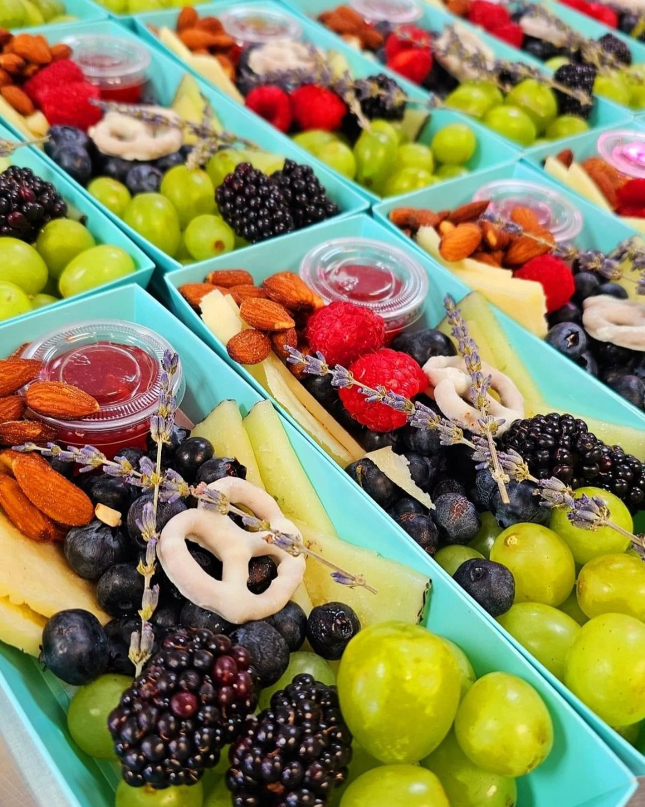 Stacked turquoise boxes filled with fresh fruit and snacks, including green grapes, blackberries, raspberries, almonds, and small clear containers of berry sauce, decorated with sprigs of lavender.