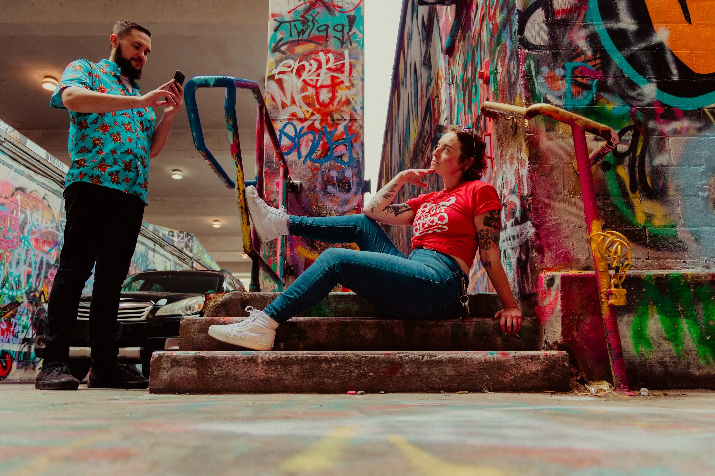 A woman with tattoos sitting on steps in front of a graffiti-covered wall, talking to a man standing nearby who is looking at his phone, under a tunnel with more graffiti.