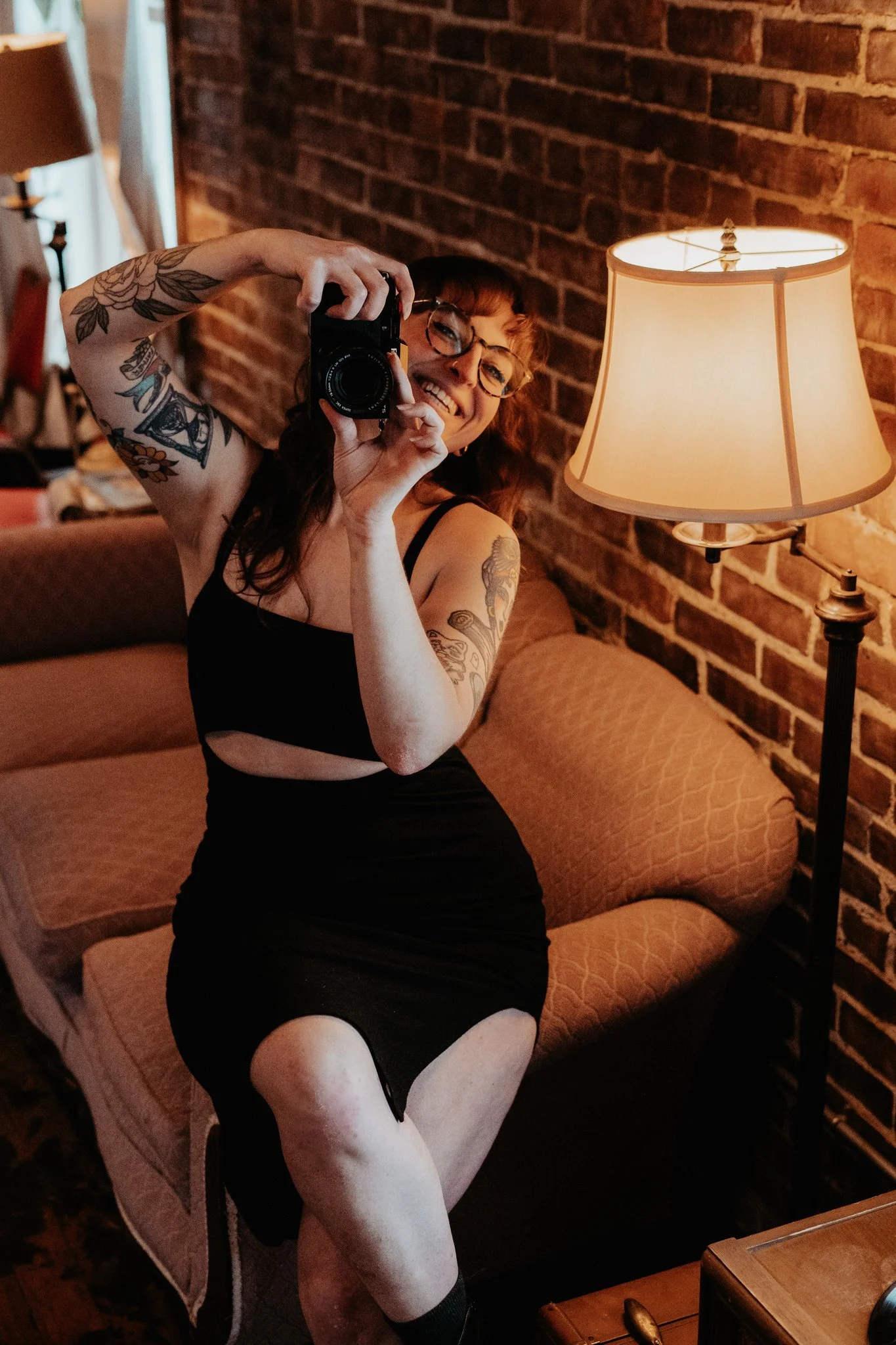 A woman with tattoos, glasses, and a black dress, smiling while taking a photo of herself in a mirror in a cozy room with a brick wall, a sofa, a floor lamp, and a wooden table.