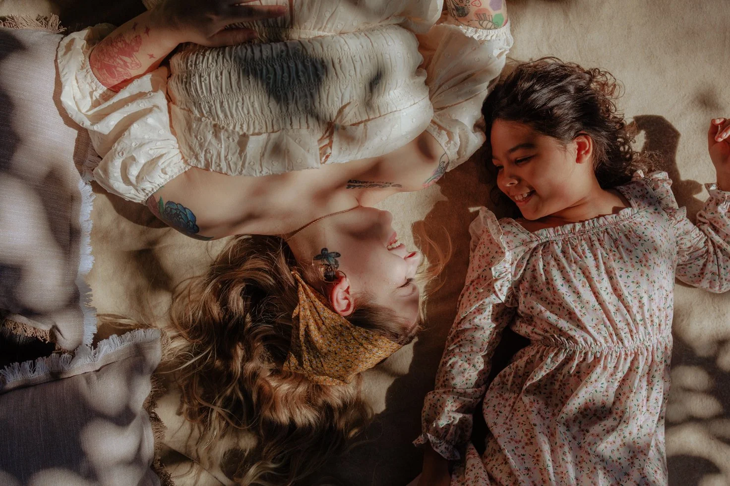 A woman and a young girl lying on their backs on a soft surface indoors, facing each other, sharing a joyful moment and smiling. The woman has tattoos and is wearing a ruffled dress with a scarf on her head, while the girl is in a floral dress with ruffles on the shoulders.