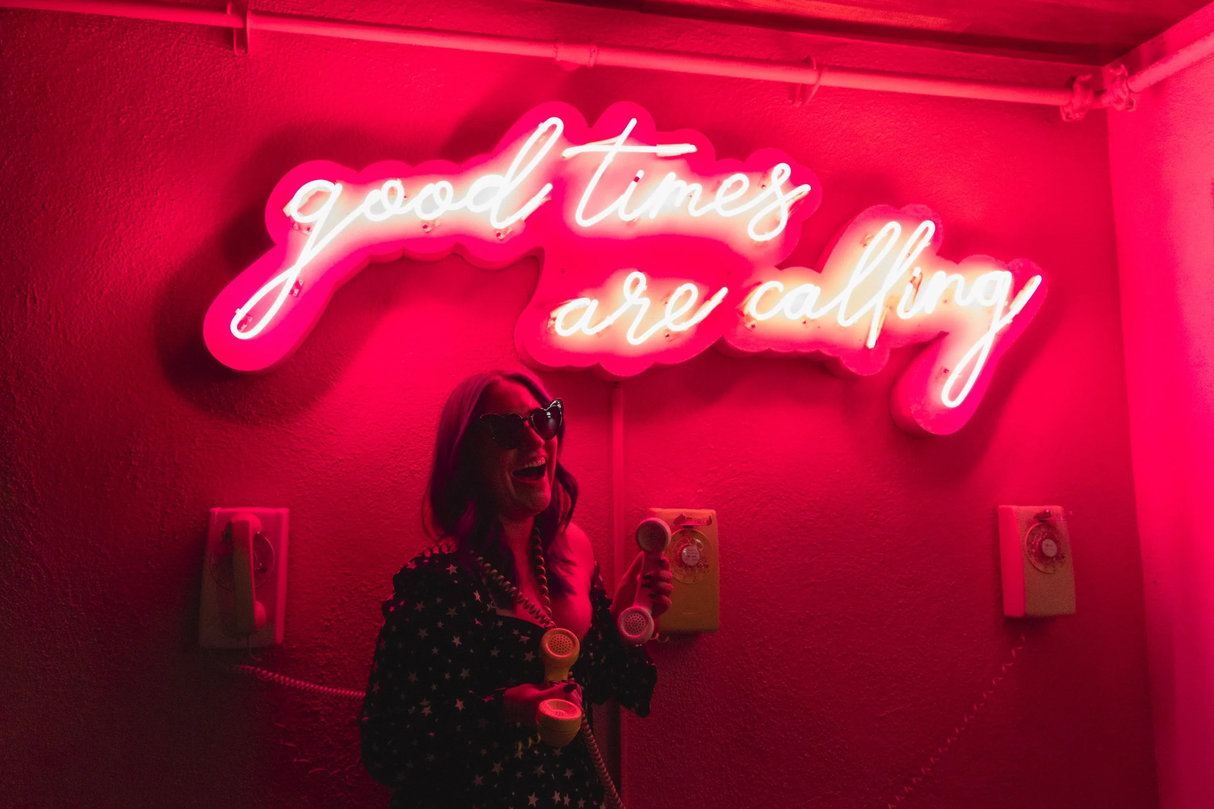 A woman wearing sunglasses and a star-patterned outfit, holding a vintage yellow telephone, standing in front of a neon pink sign that reads 'good times are calling' on a red wall.