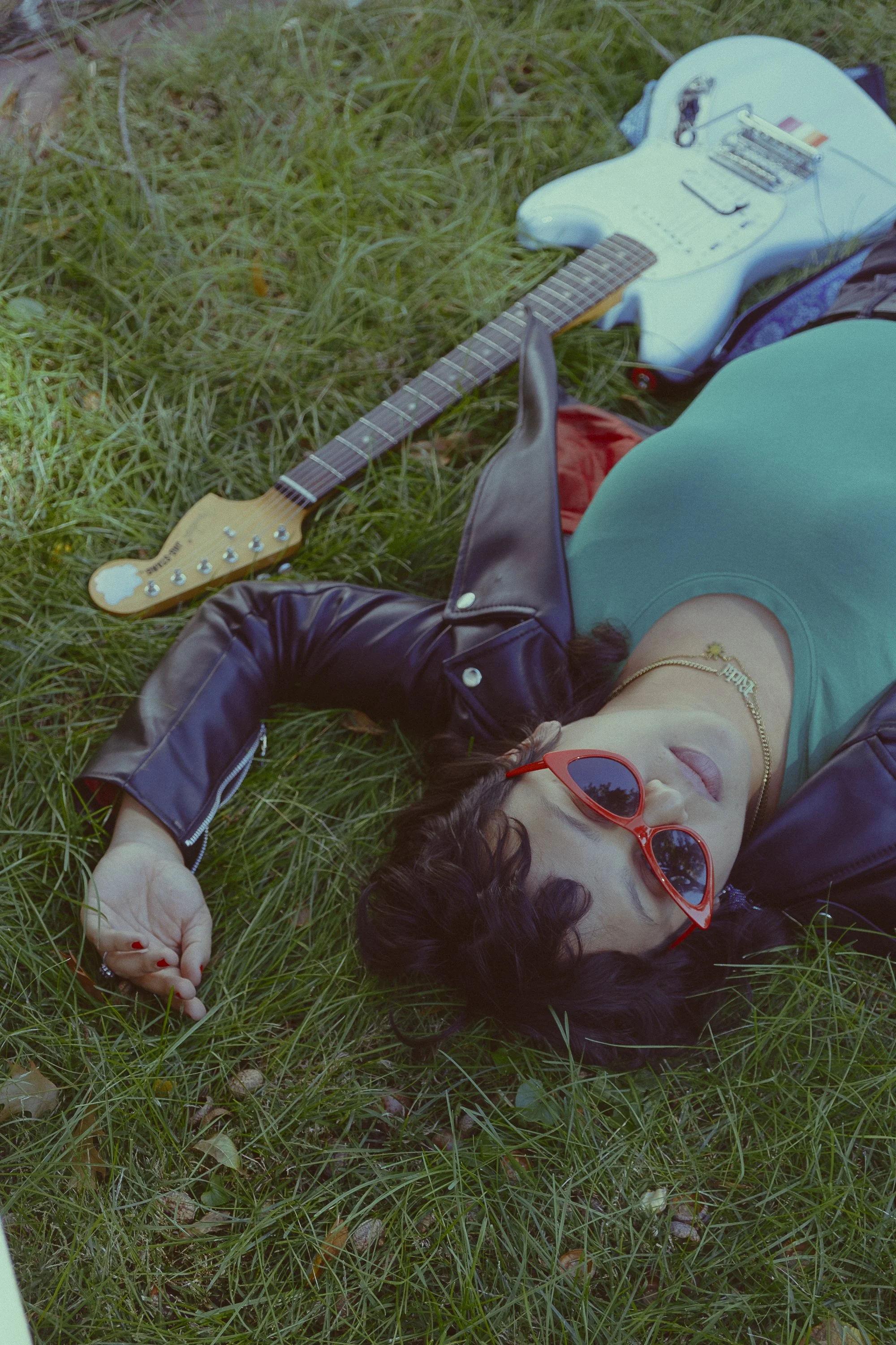 Musician promo portrait RIcki Marvelous laying on grass with guitar, leather jacket and red sunglasses