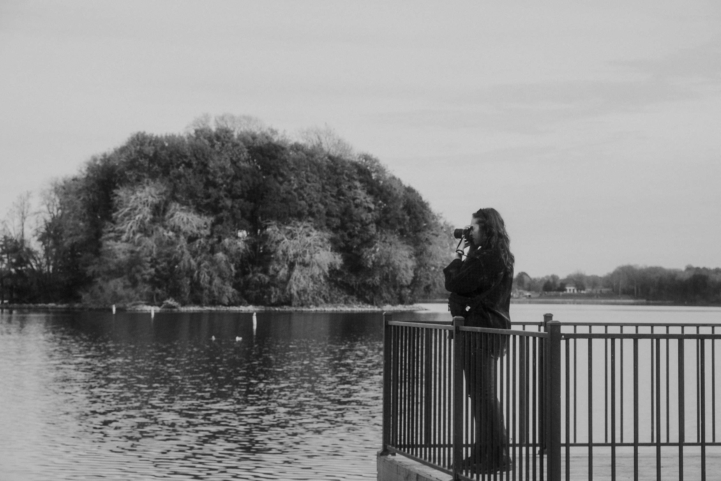 A woman with long hair taking a photograph with a camera on a lakeside deck in black and white.