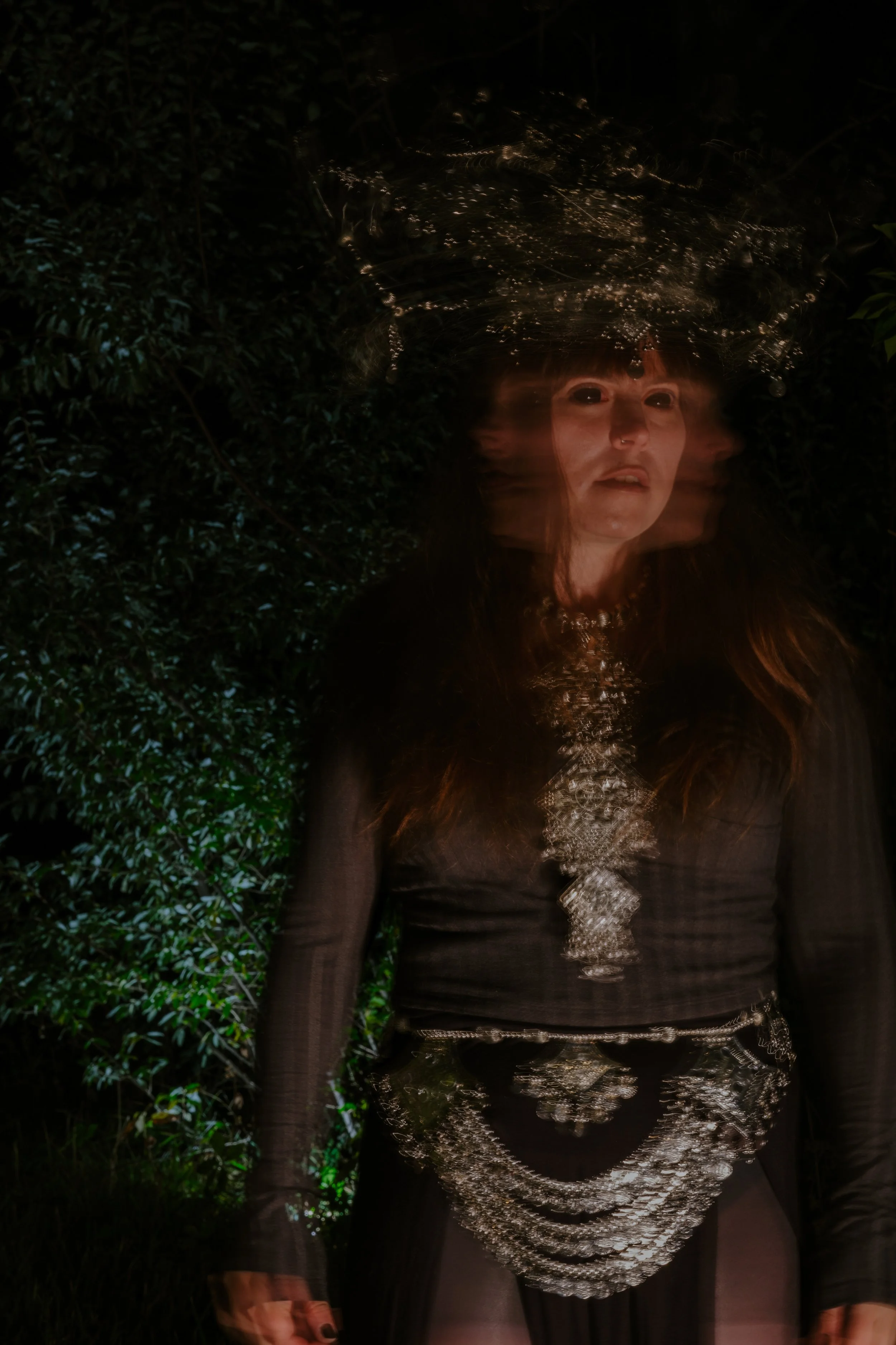 A woman with long hair, dressed in black with silver jewelry, standing outdoors at night with dark, leafy background, and a spider web above her head.