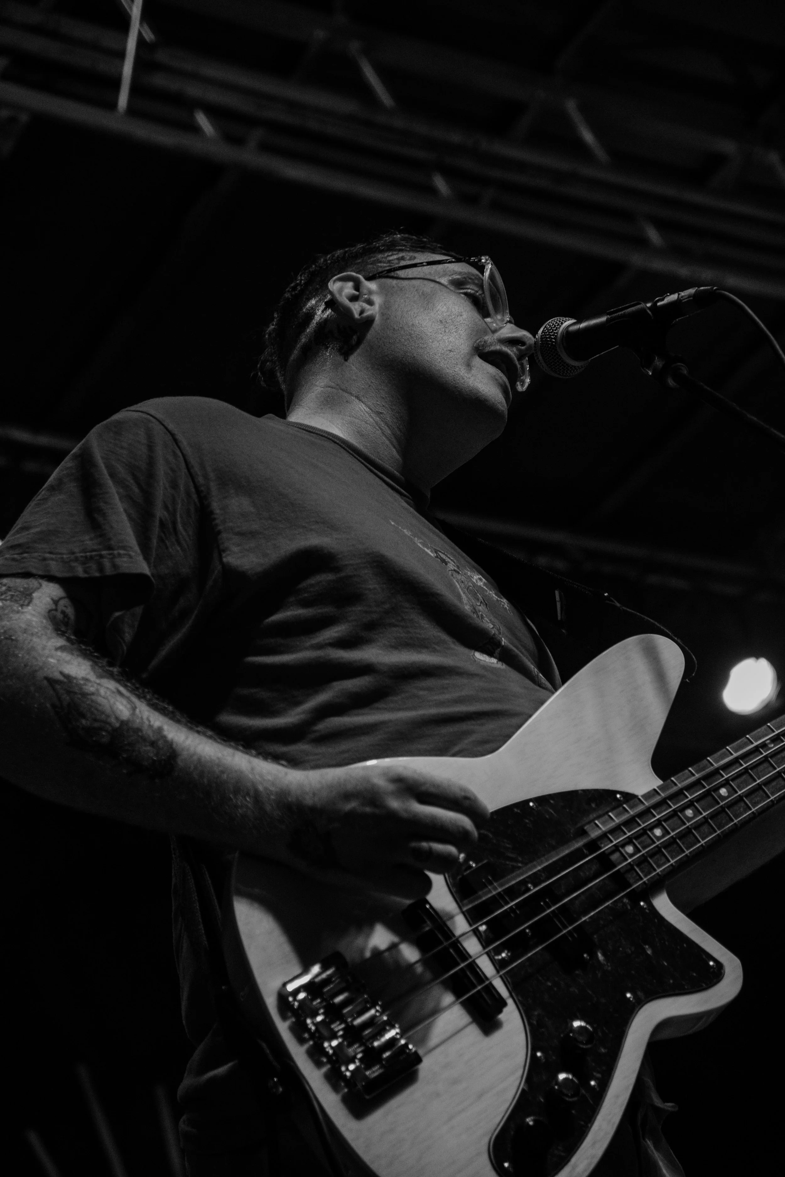 A man with tattoos, glasses, and a headset singing into a microphone while playing an electric bass guitar on stage.