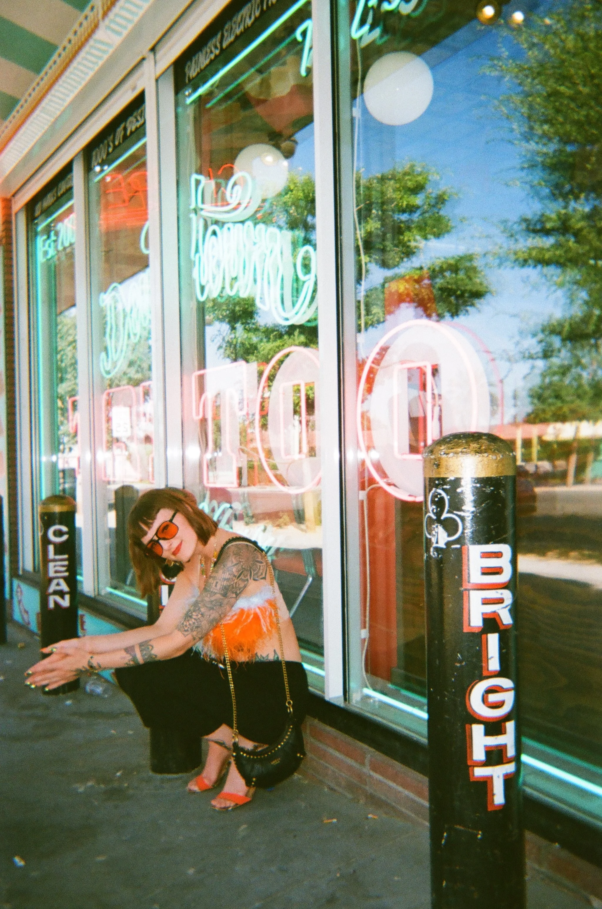 A woman with tattoos, wearing sunglasses, an orange top, black pants, and orange high heels, squatting on the sidewalk outside a boutique with neon signs that say 'Open' and 'Closing,' reflected in the windows.