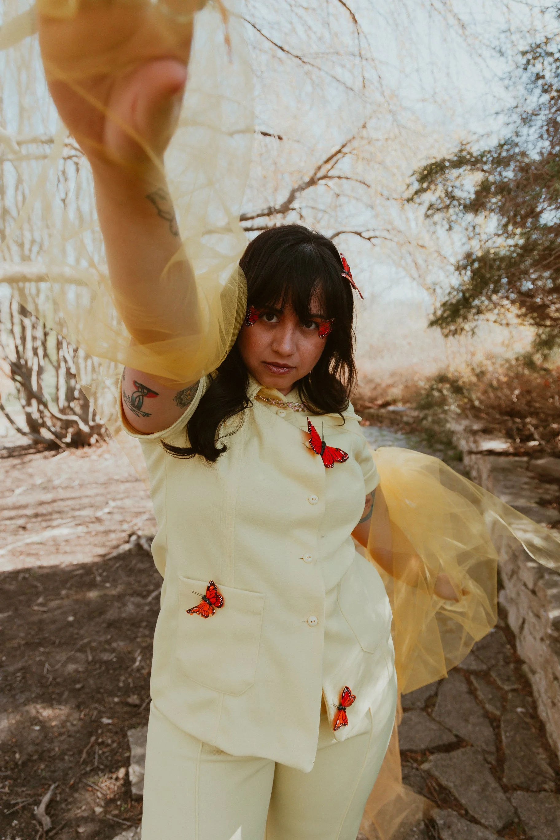 A woman with dark hair and tattoos on her arm is standing outdoors in a wooded area, wearing a pale yellow outfit decorated with red and orange butterfly embellishments, and yellow tulle accents. She is reaching out towards the camera with her hand, with a serious expression on her face.