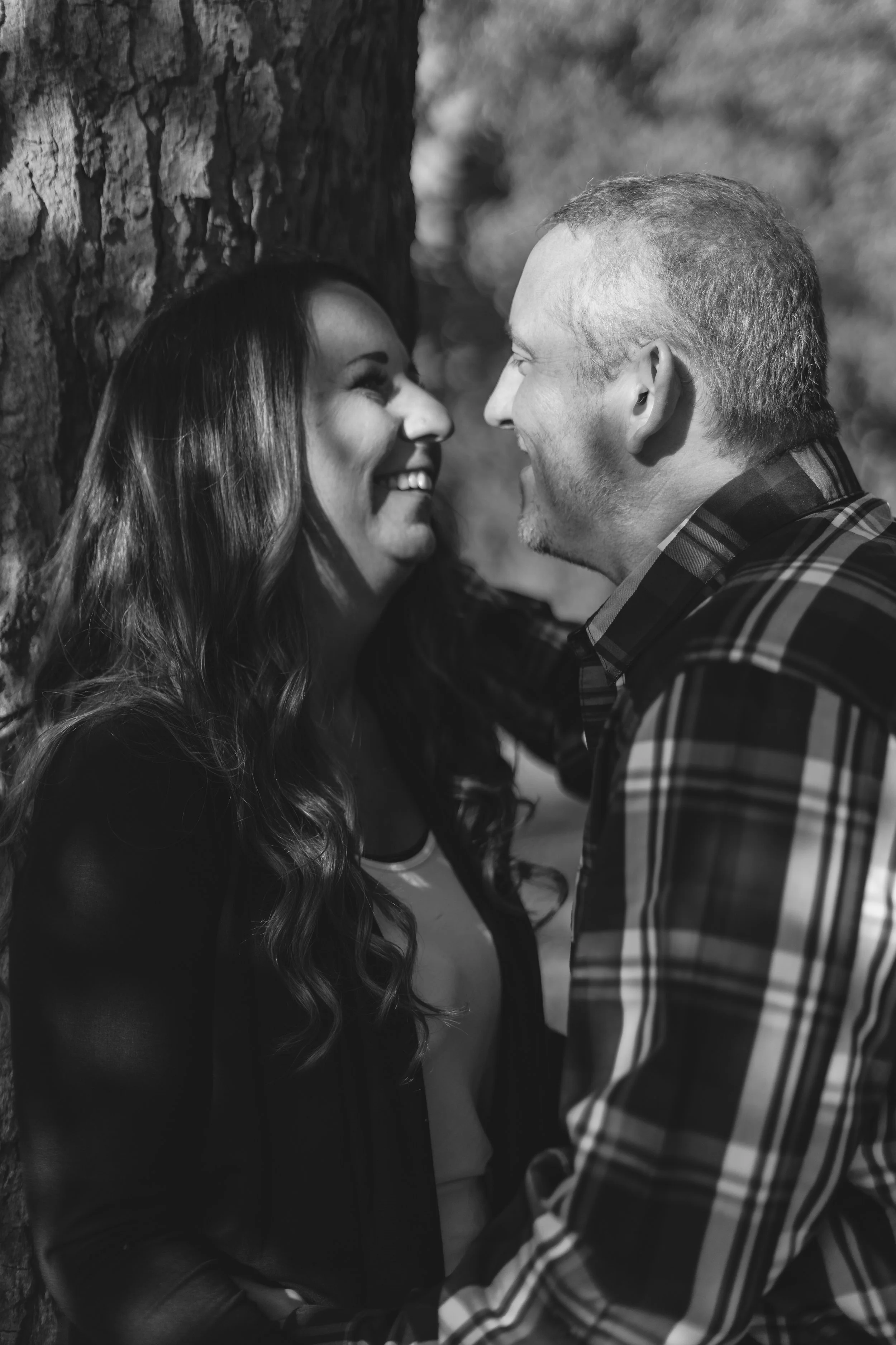 A black and white photograph of a man and woman smiling at each other outdoors near a tree. The woman has long wavy hair, and the man has short hair and a beard. They are leaning close to each other, appearing happy and affectionate.