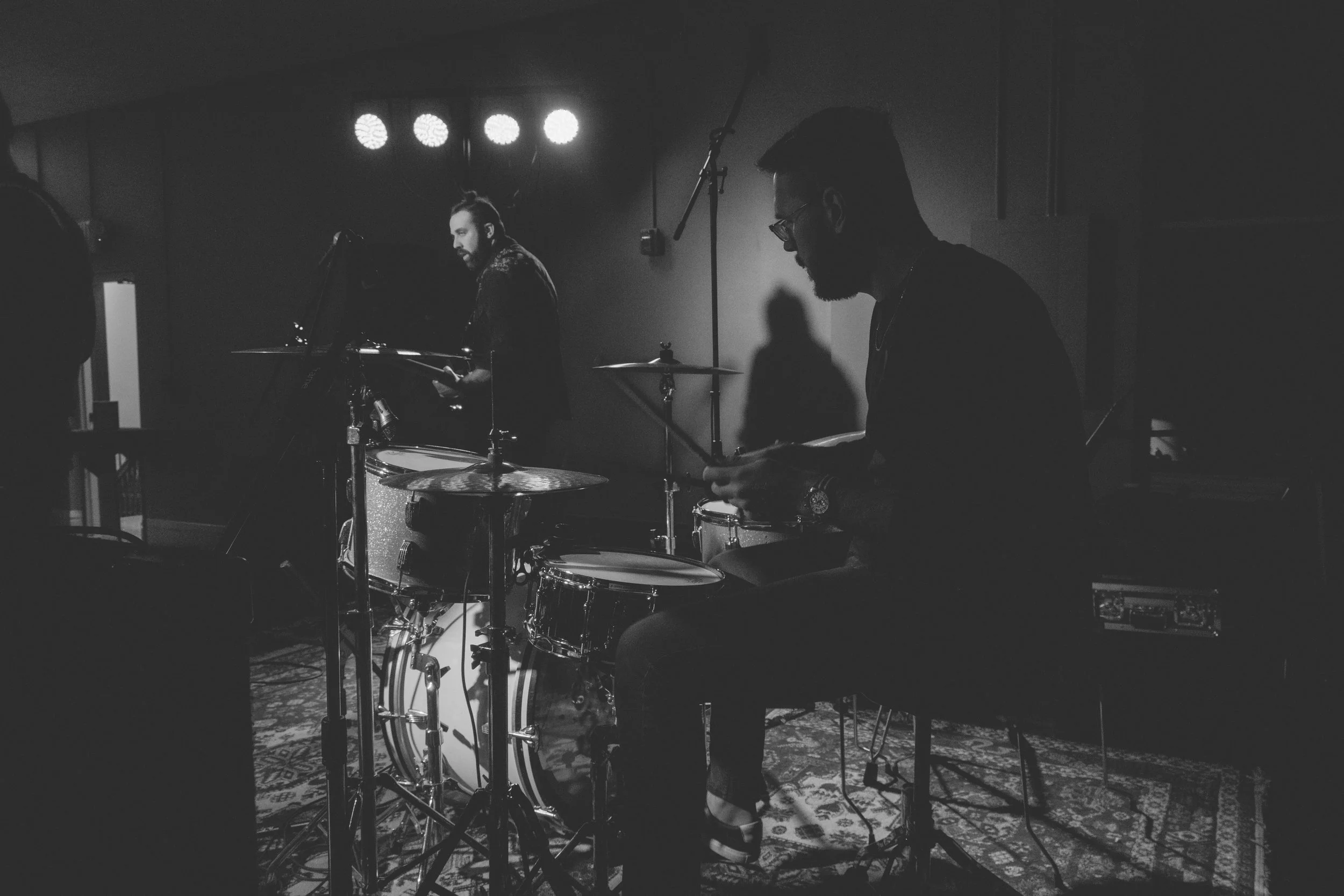 Two musicians playing drums and percussion during a live performance on a stage with spotlighting.