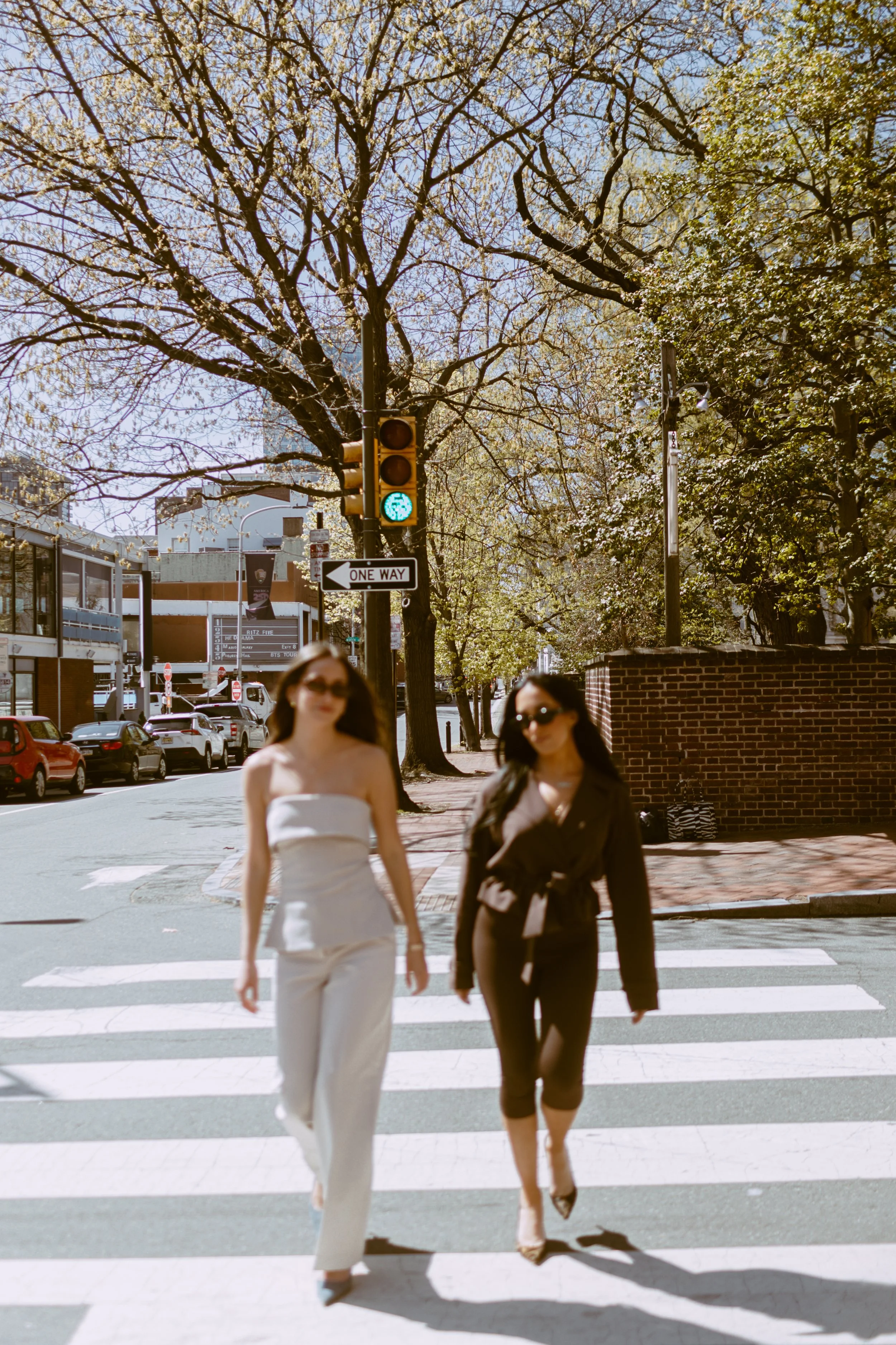 Two women walking on a city street crosswalk during daytime, with trees and buildings in the background, and traffic lights showing a green pedestrian signal. Byob Marketing Philadelphia Social Media Management Content Creation