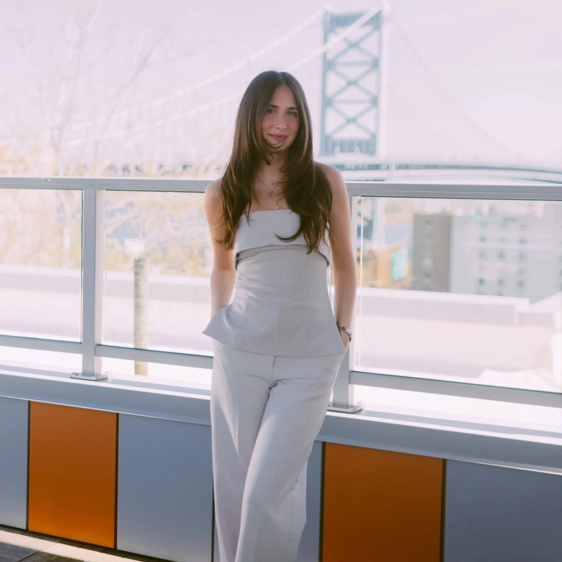 A woman with long brown hair, wearing a strapless white dress, is standing on a balcony with a view of a cityscape. Byob Marketing Philadelphia Social Media Management Content Creation