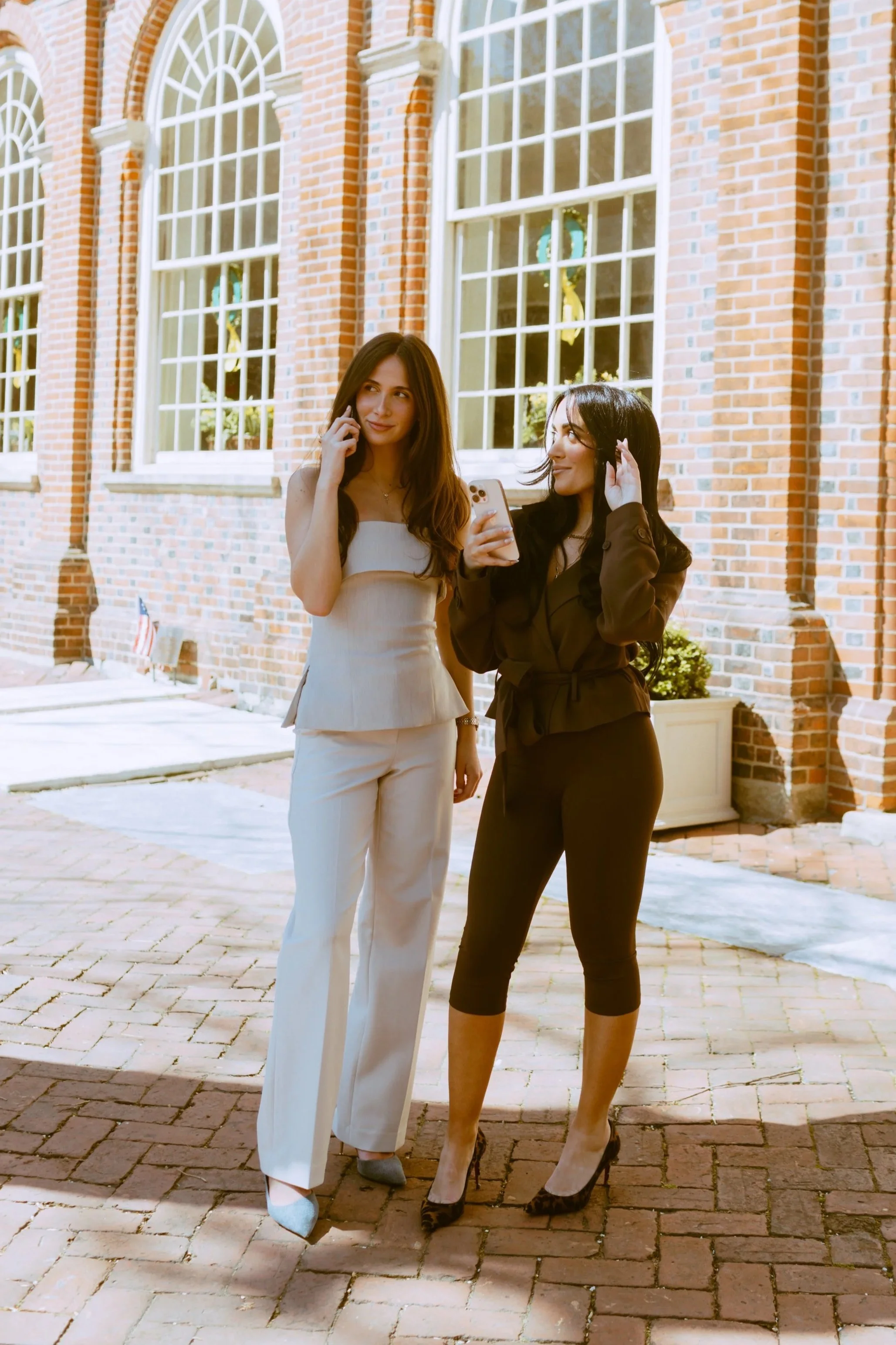 Two women standing outside in front of a brick building with large windows. One woman is on the phone and the other is holding a smartphone. Byob Marketing Philadelphia Social Media Management Content Creation