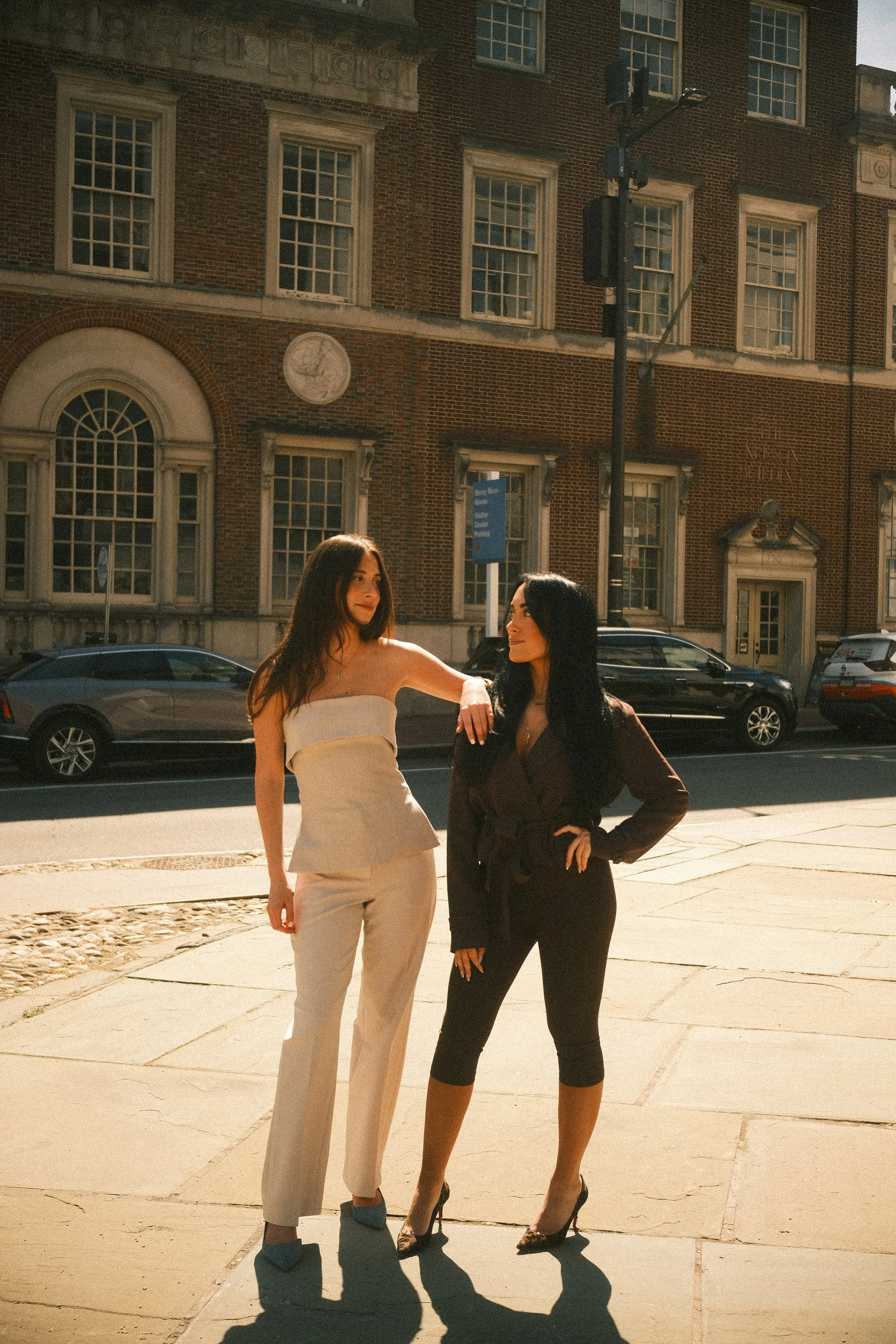 Two women standing on a city sidewalk engaging in conversation, with vintage brick buildings and parked cars in the background during daytime. Byob Marketing Philadelphia Social Media Management Content Creation