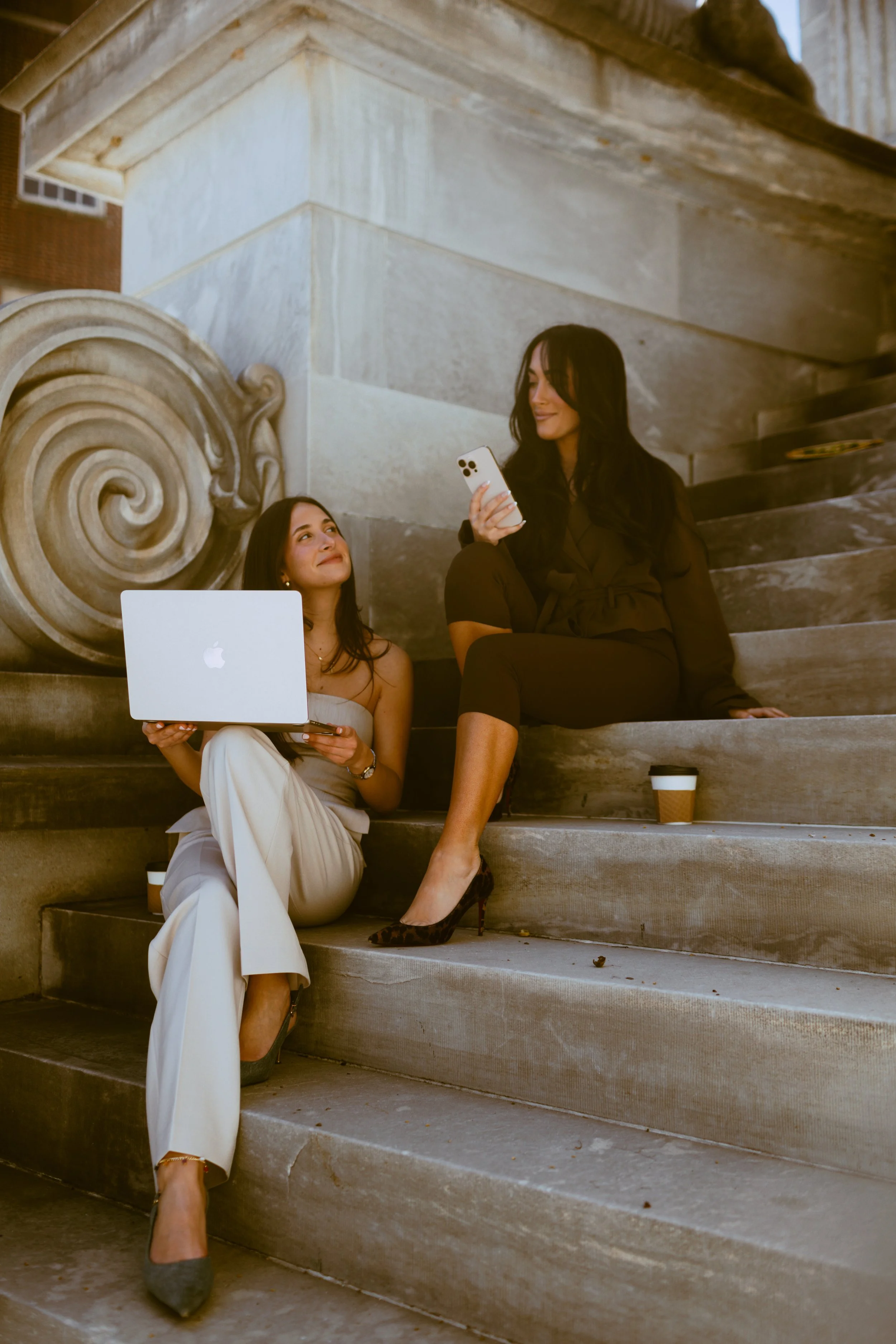 Two women sitting on stone steps in front of a building, with one holding a laptop and the other looking at her phone. Coffee cups are on the steps. Byob Marketing Philadelphia Social Media Management Content Creation