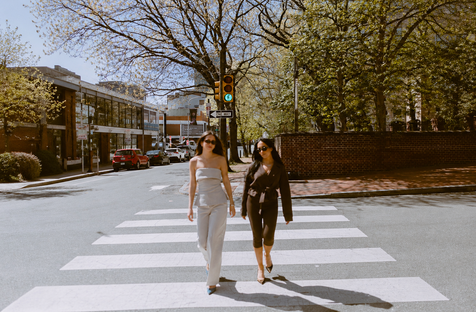 Two women walking on a crosswalk in an urban area on a sunny day, with trees, cars, and buildings in the background. Byob Marketing Philadelphia Social Media Management Content Creation
