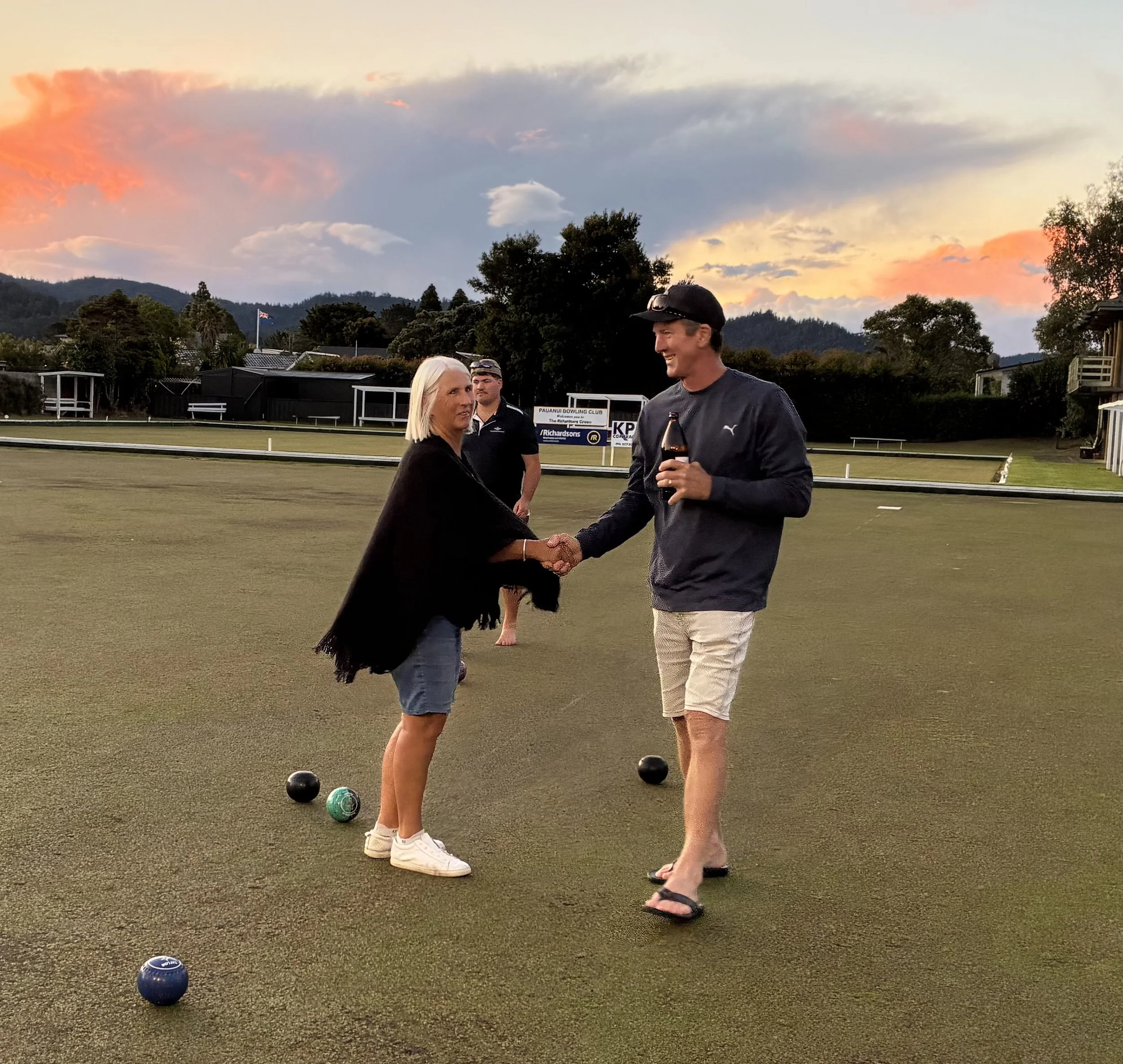 Two women shaking hands on a lawn bowls green with three bowls on the ground, set against a sunset sky with colorful clouds. One woman has blonde hair and is wearing a black shirt, while the other has short dark hair, sunglasses on her head, and is w