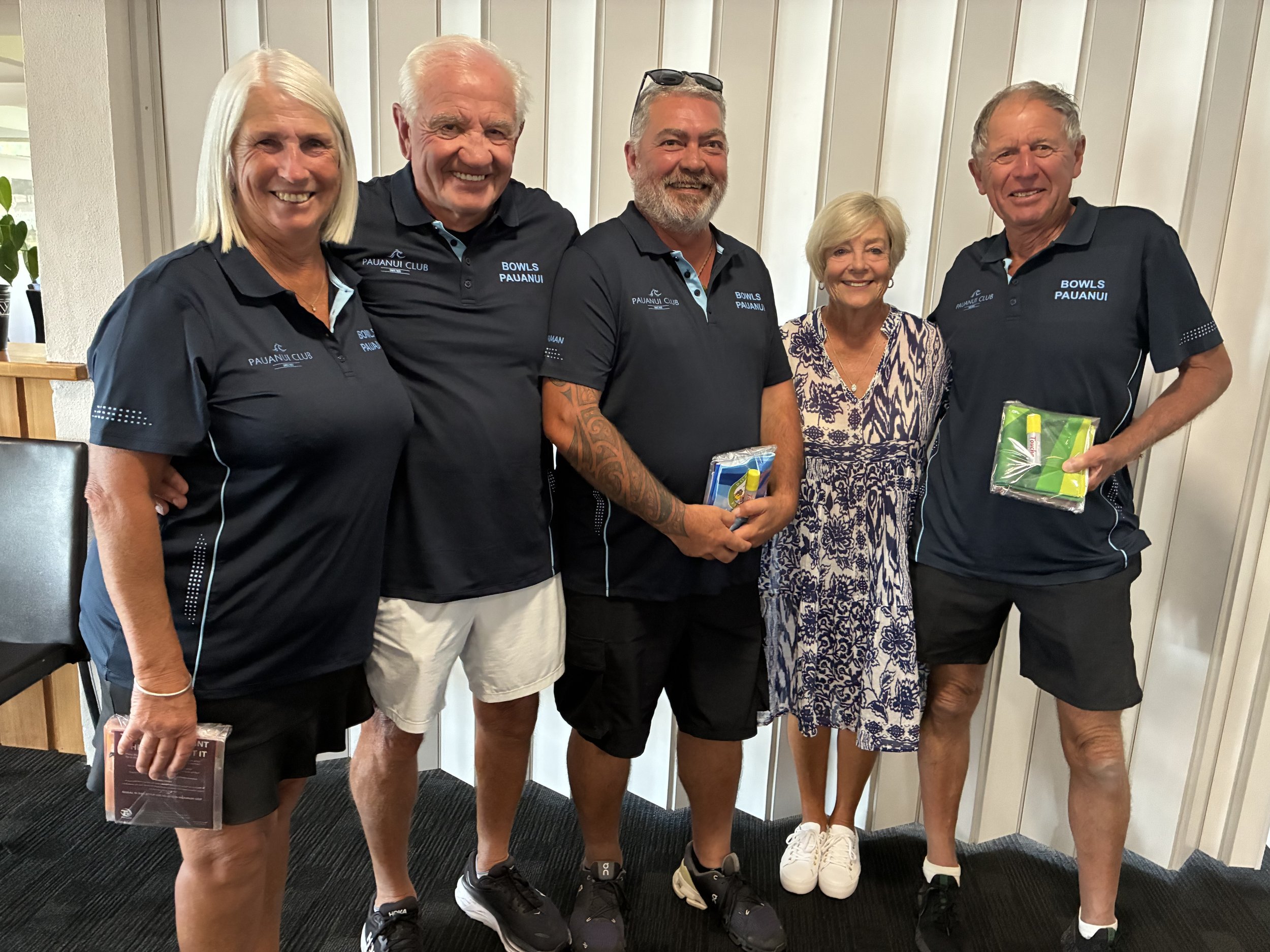 A group of five smiling adults, three men and two women, standing together indoors. They are wearing matching navy blue shirts with the words 'BOWLS PAUANUI' embroidered on the left side. The woman on the far right is holding a package of tissues, an